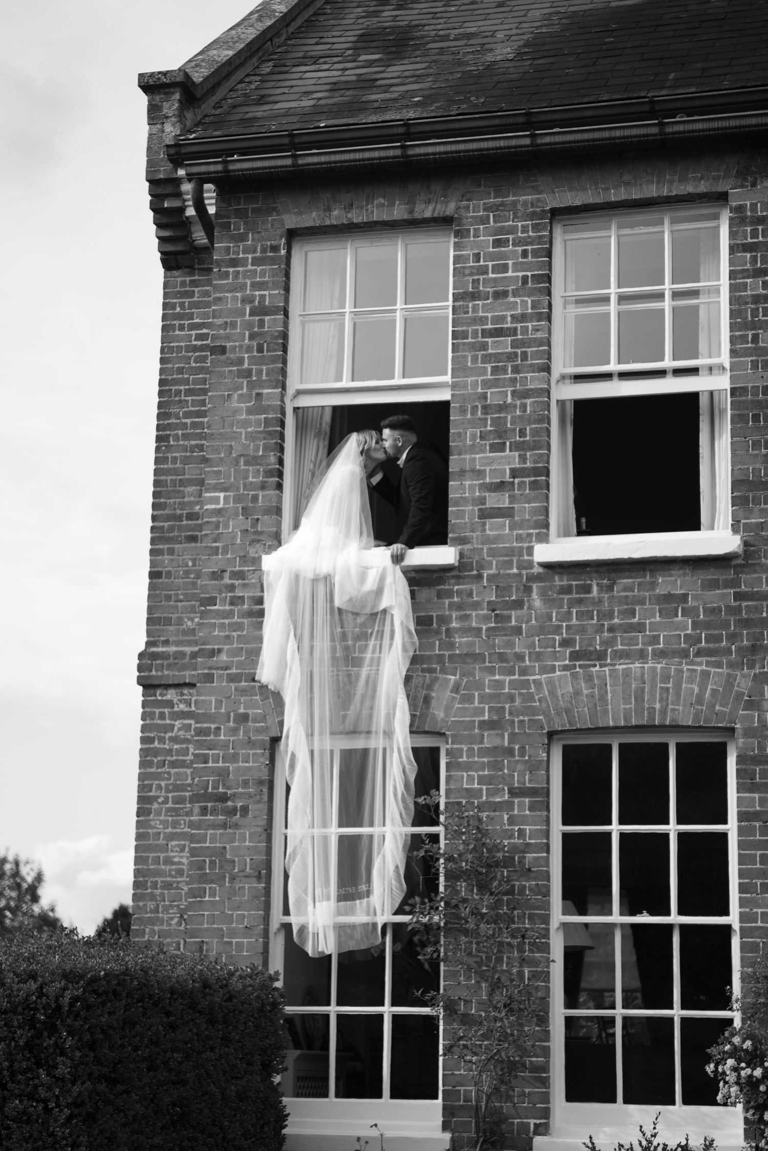 Black and white image of newlyweds sharing a kiss in an open window with the bride's veil cascading down.