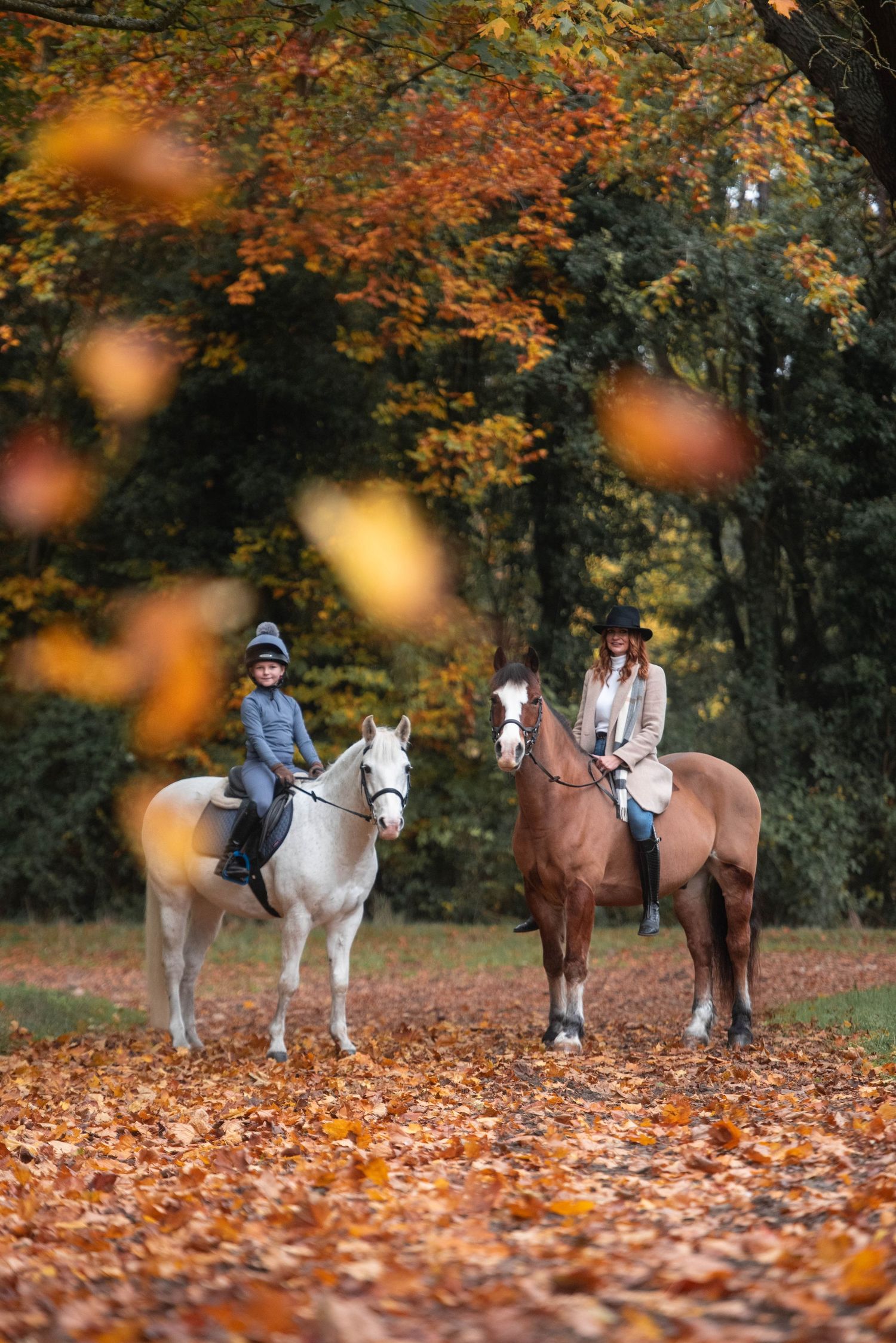 Two riders on white and brown horses in an autumn forest with colorful fallen leaves.