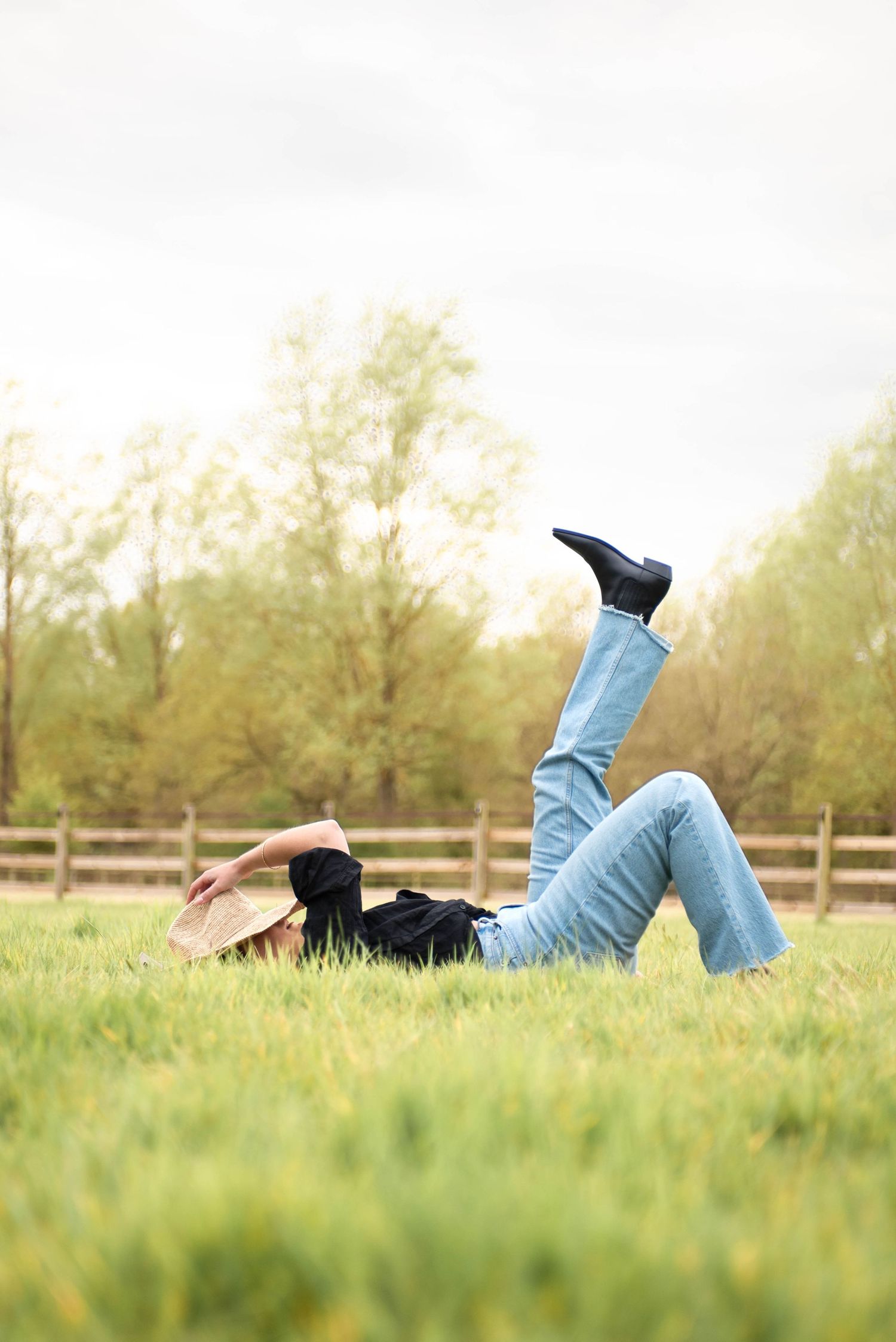 Woman in jeans and cowboy boots lies on green grass with legs raised in outdoor field setting.