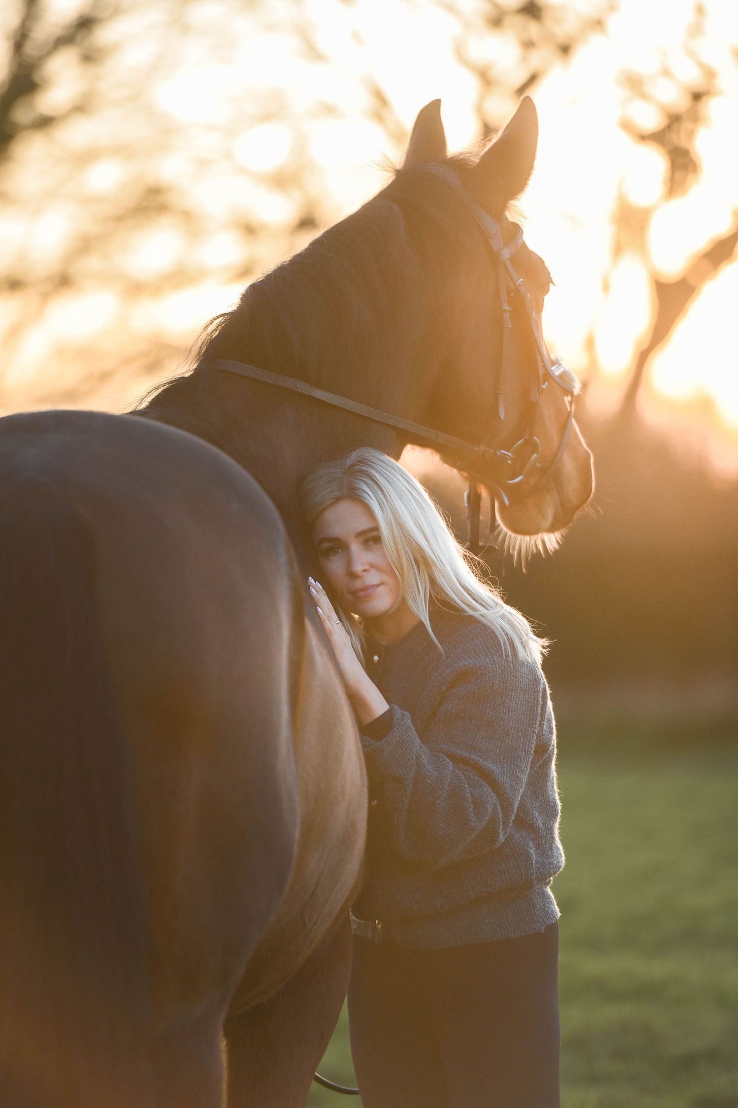 Blonde woman standing close to a bay horse, leaning against it affectionately in golden sunset light.