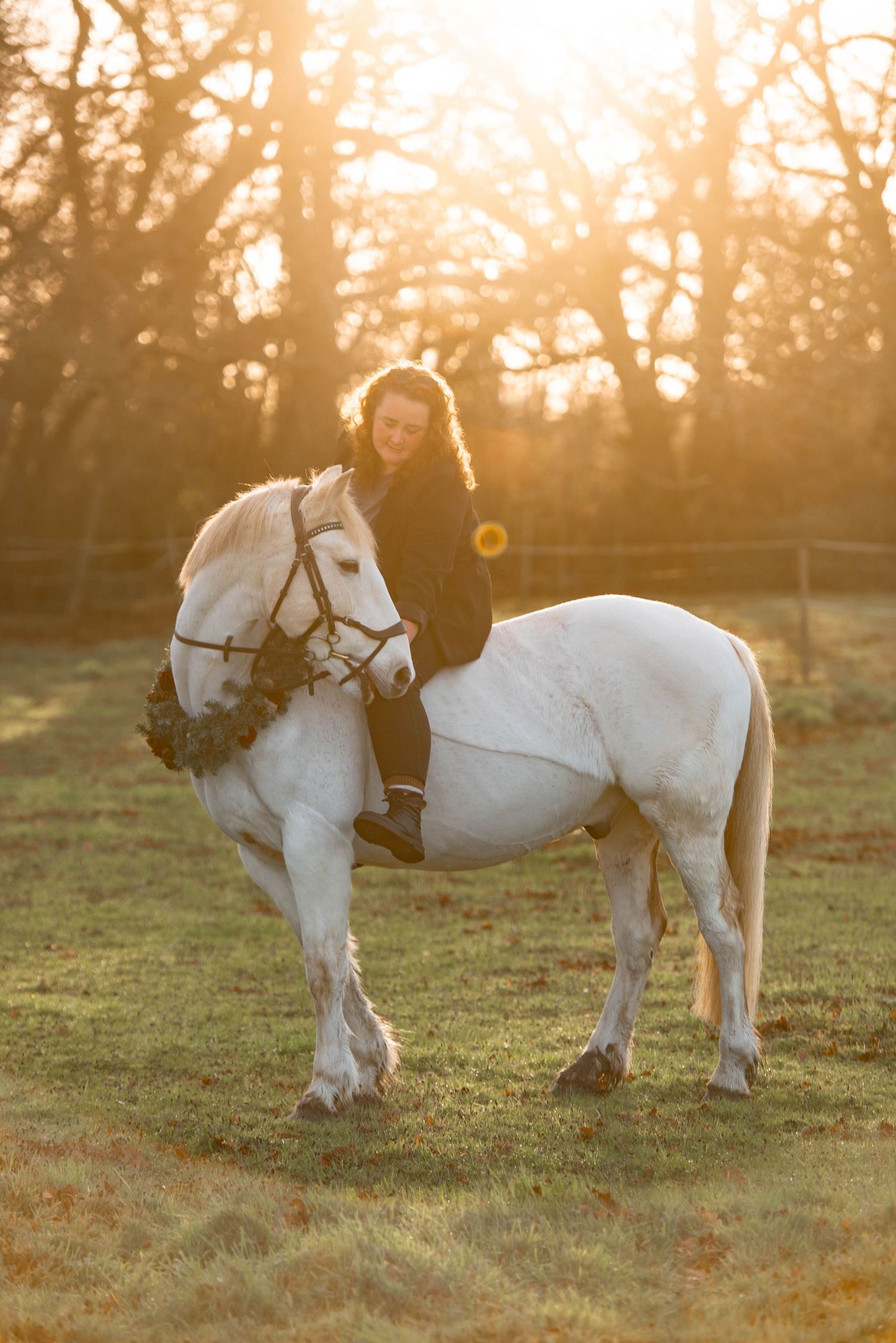 A smiling woman rides a white horse bareback in a sun-drenched field during golden hour.