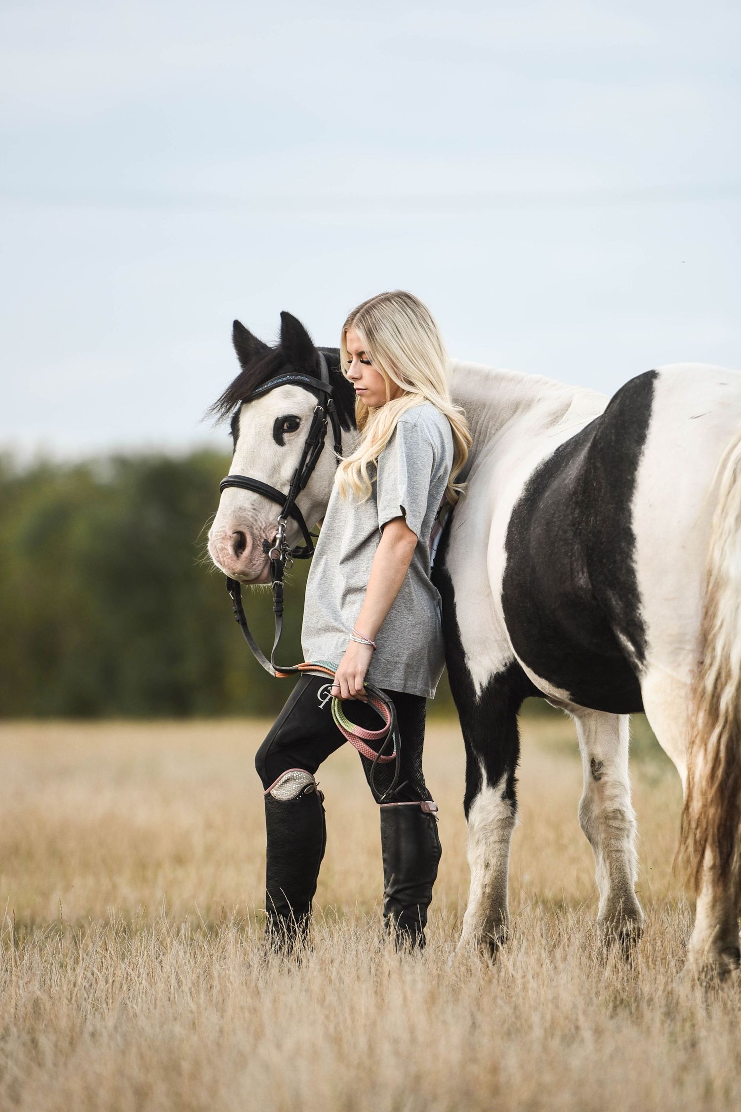Blonde woman in grey shirt leaning against black and white paint horse in field.