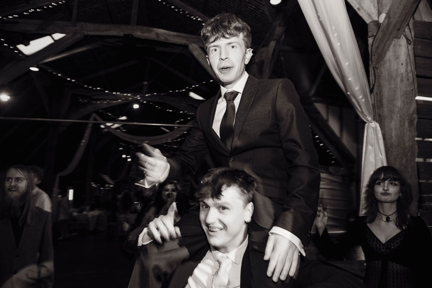 Black and white photo of two men at a wedding reception, one sitting on the other's shoulders, laughing.