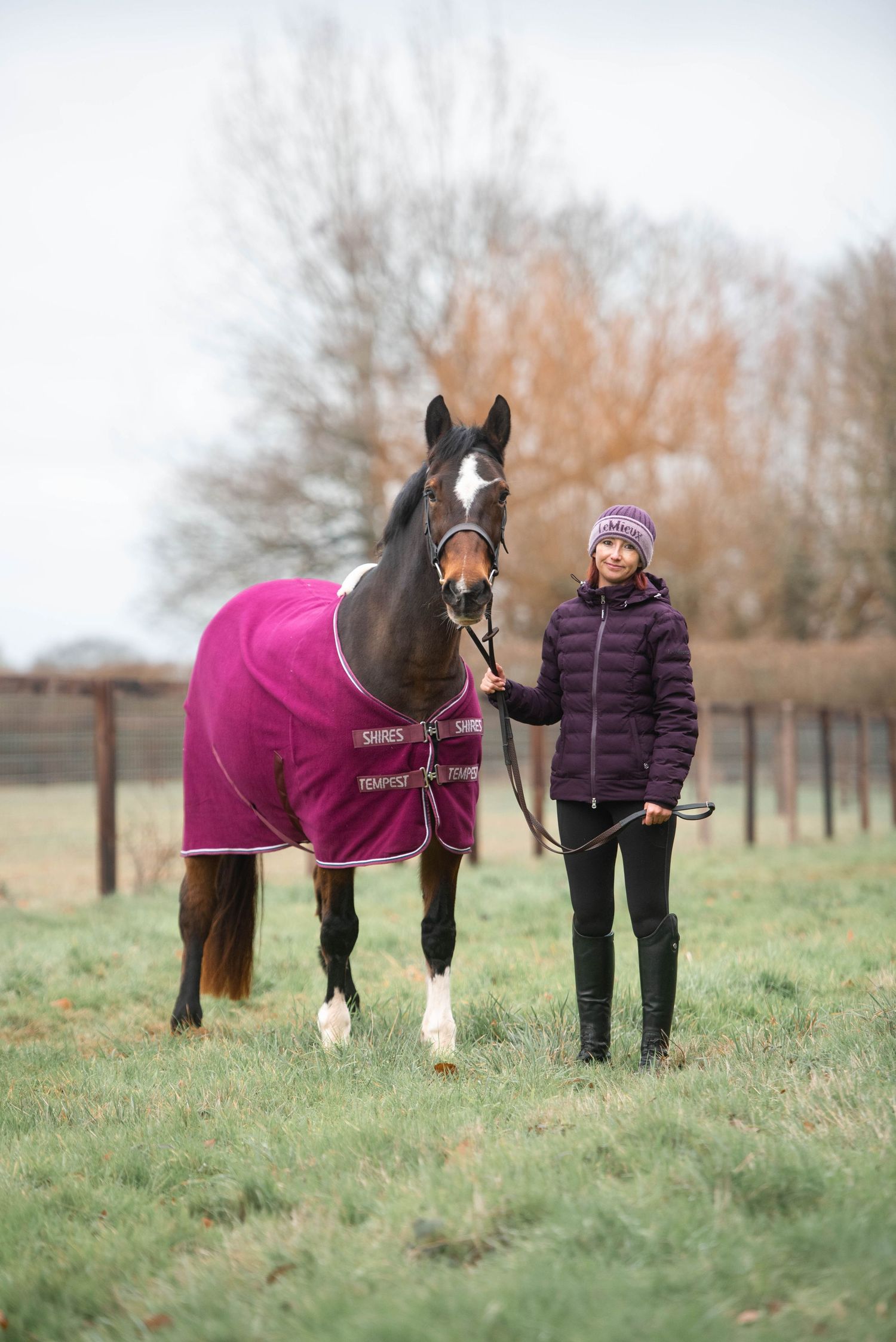 Girl in purple hat and jacket standing with a tall dark horse in a purple rug in a field.