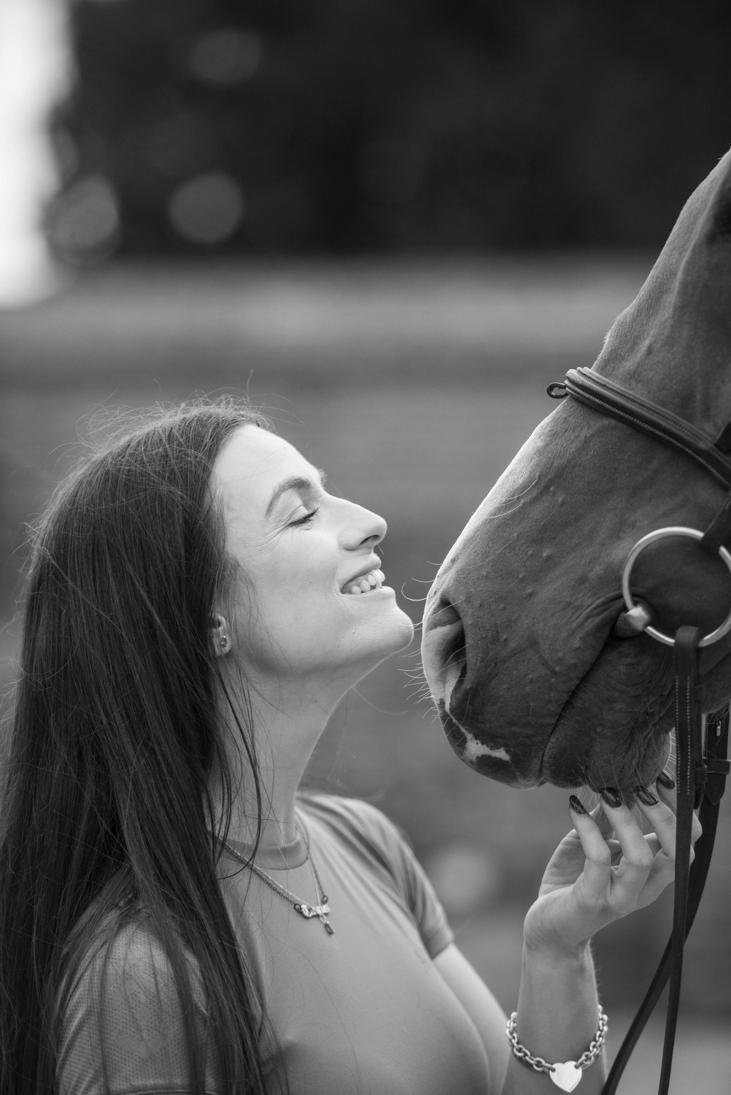 Black and white portrait of a smiling woman with long hair sharing a tender moment with a horse.