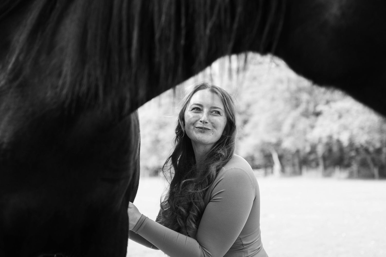Black and white photo of a smiling woman with long hair standing beside a dark horse outdoors.
