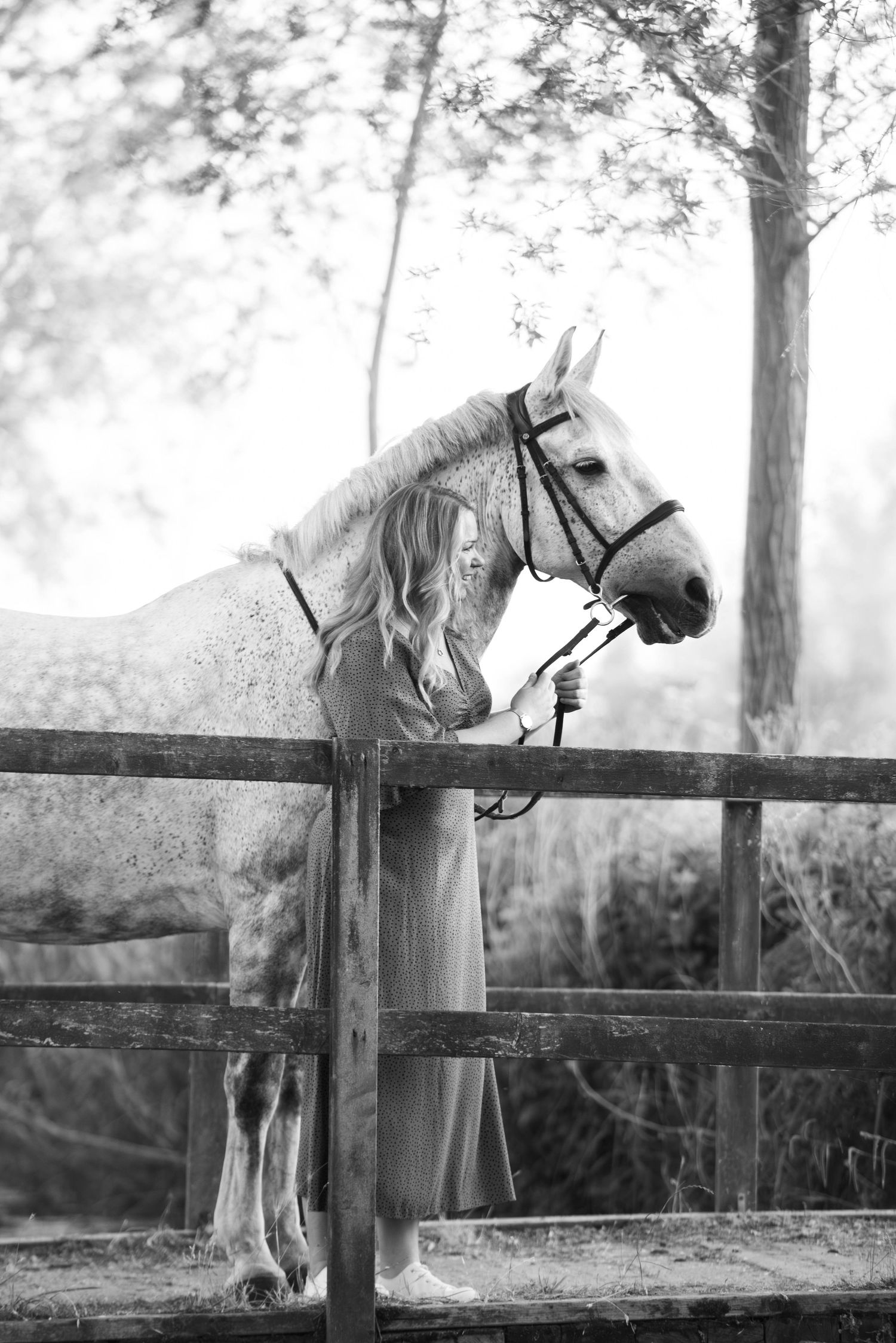 Black and white photo of a woman in a gown with a white horse at a wooden fence.