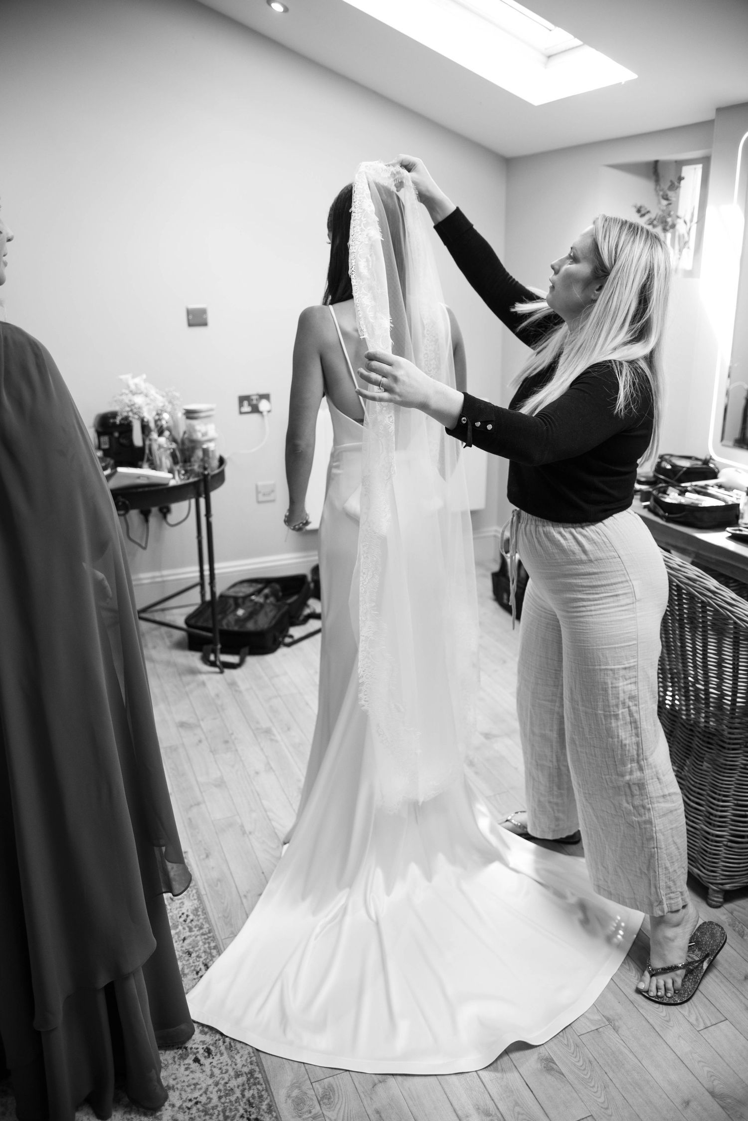 Black and white photo of a bride having her wedding veil adjusted by a blonde woman in a bright dressing room.
