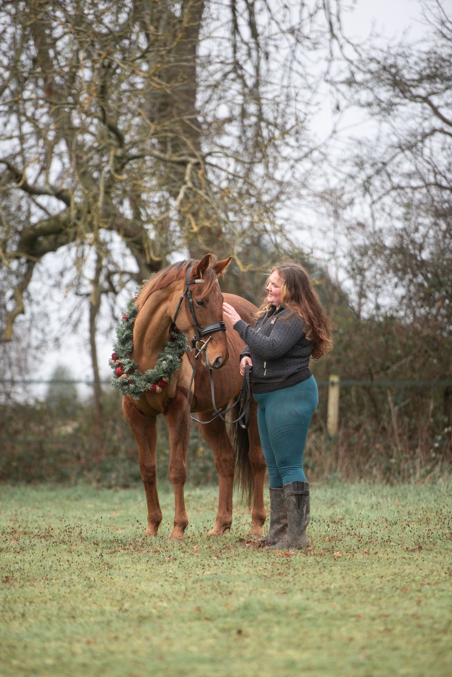 Woman with long brown hair standing beside a chestnut horse adorned with a Christmas wreath outdoors.