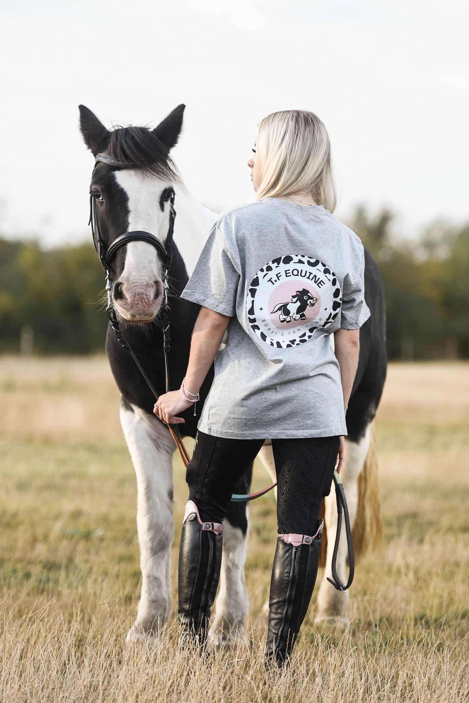 Blonde woman in TF Equine grey tee standing with black and white horse in field.