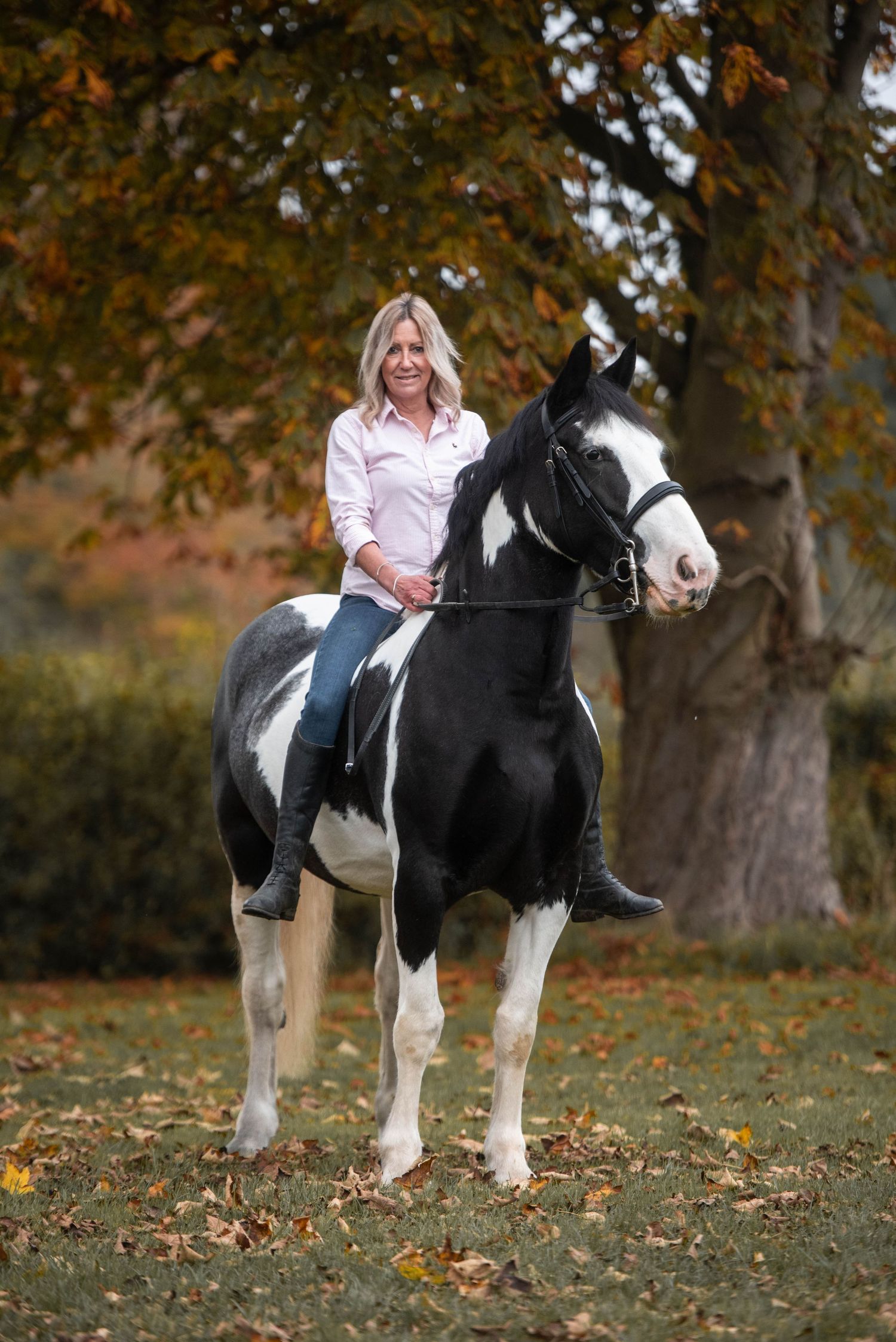 Blonde woman in pink shirt riding a black and white paint horse among autumn foliage.