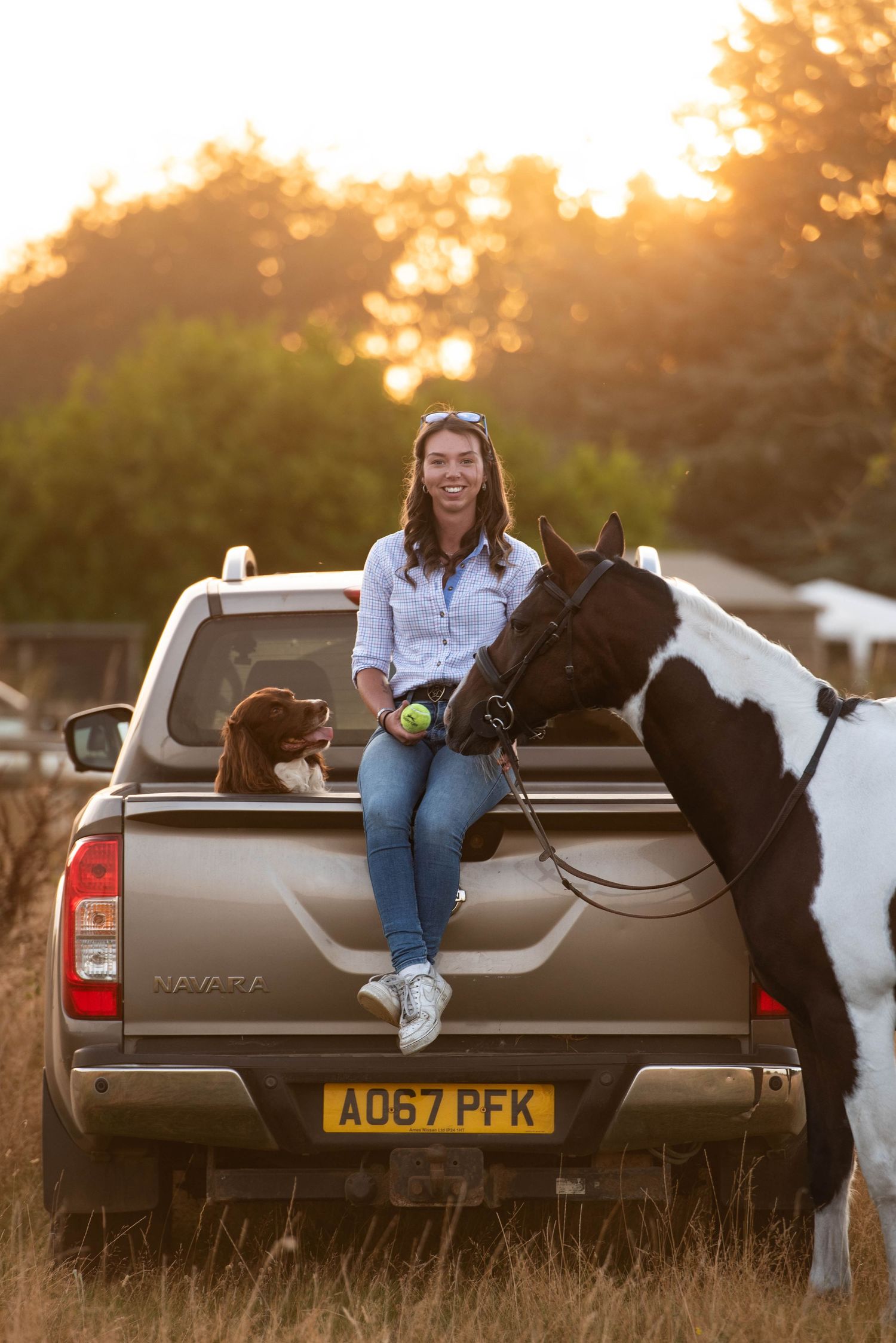 A smiling woman sits on a truck tailgate at sunset with a dog and a black and white horse.