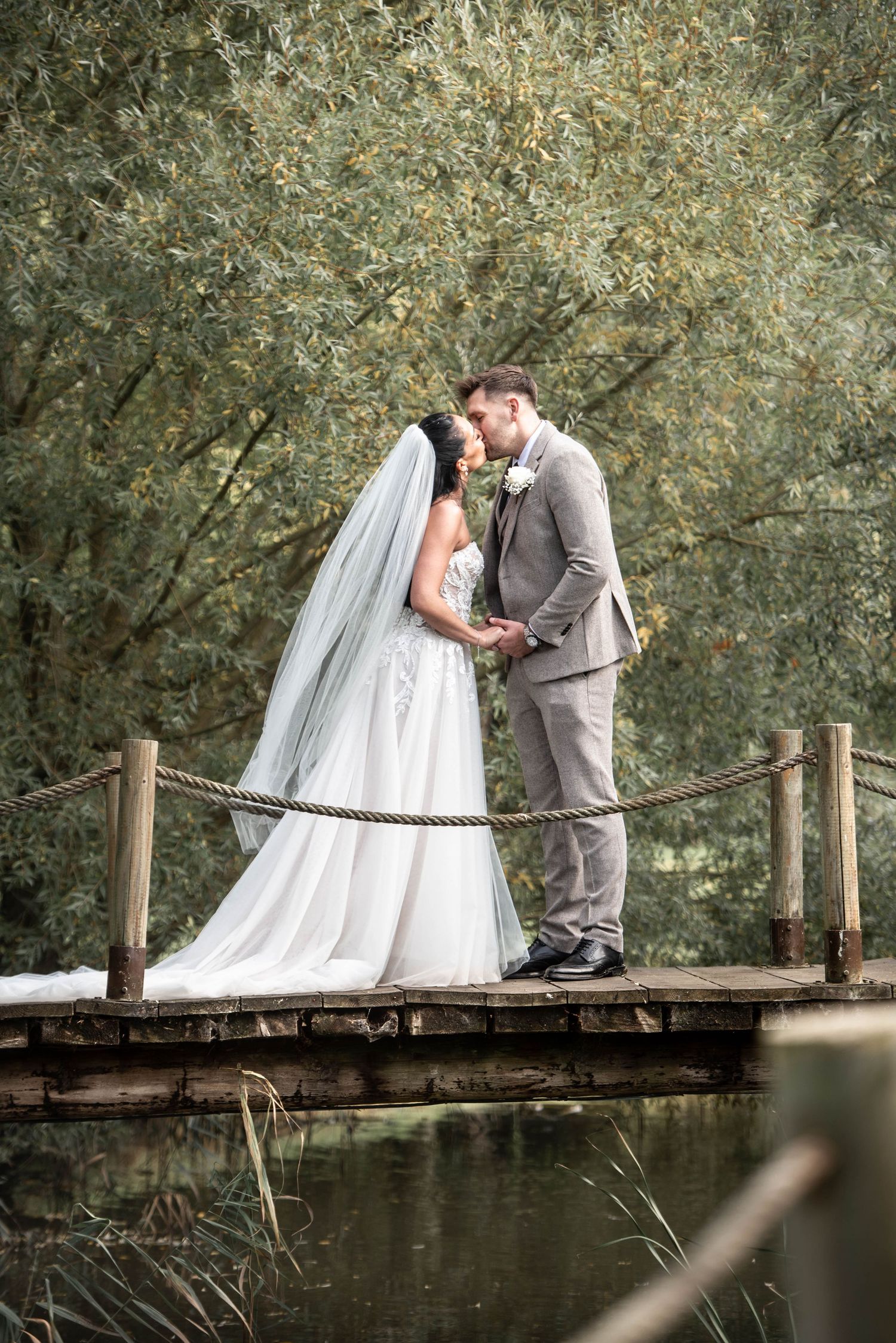 Bride and groom sharing a romantic kiss on a wooden bridge over water surrounded by lush greenery.