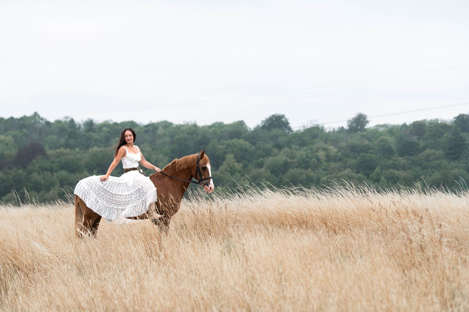Woman in white dress riding a chestnut horse through tall golden grass fields with green trees in background.