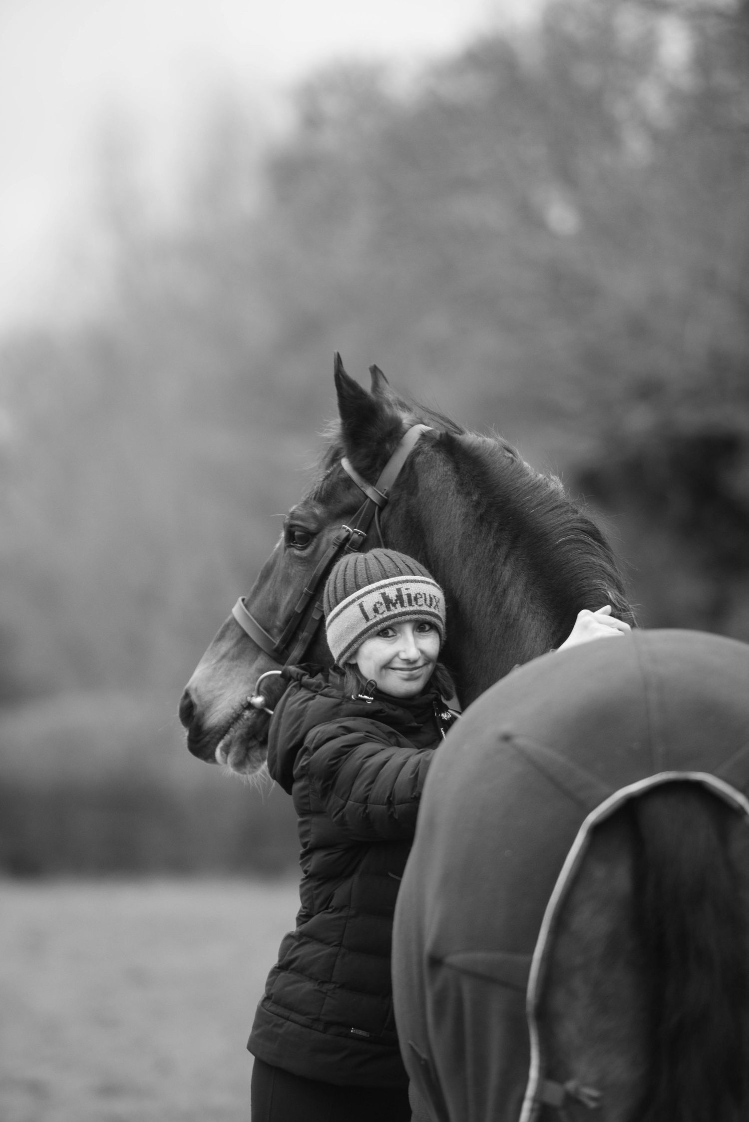 Black and white photo of a smiling girl in a LeMieux hat embracing a dark horse outdoors.