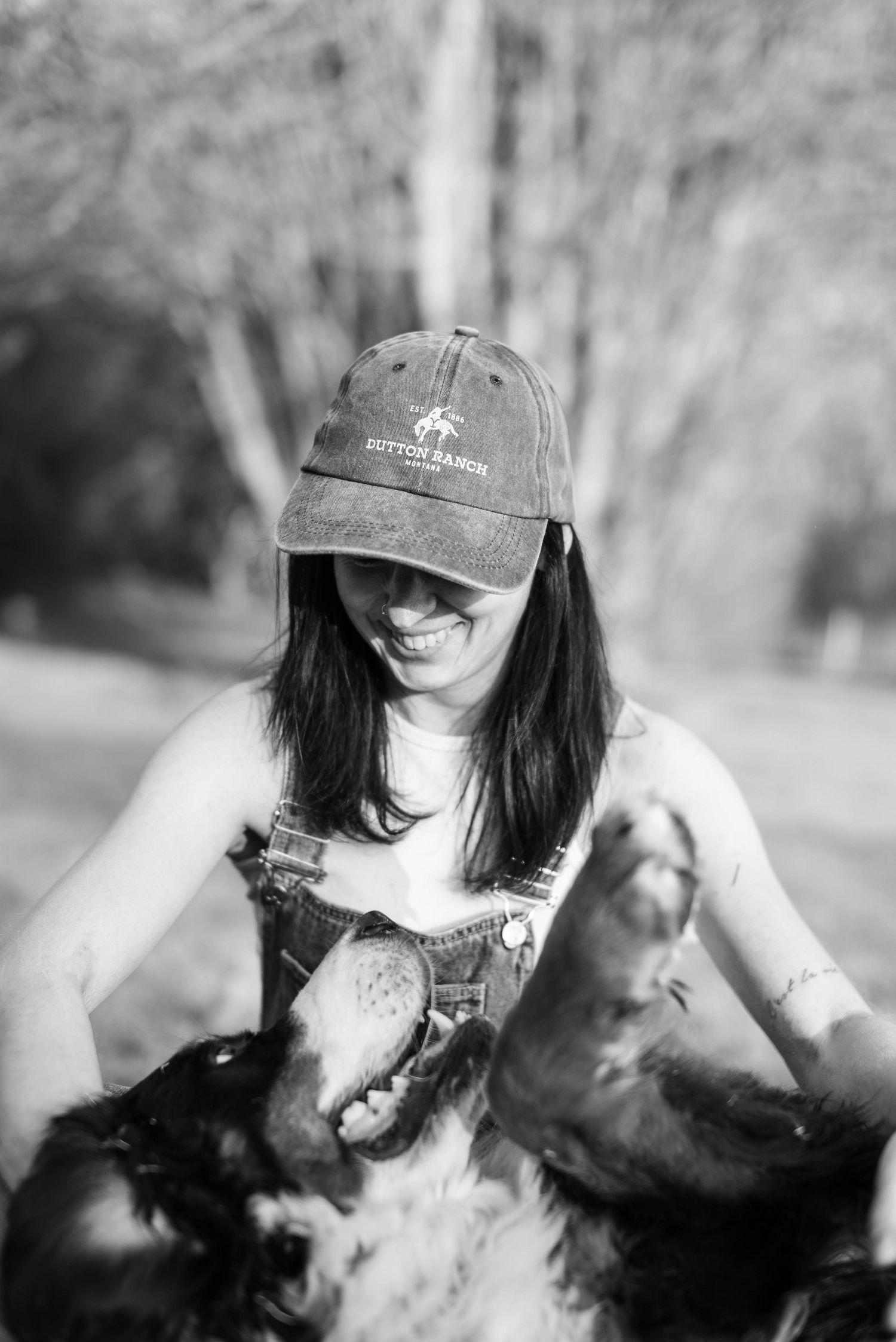 A smiling woman in overalls and cap playing with a dog outdoors in black and white.