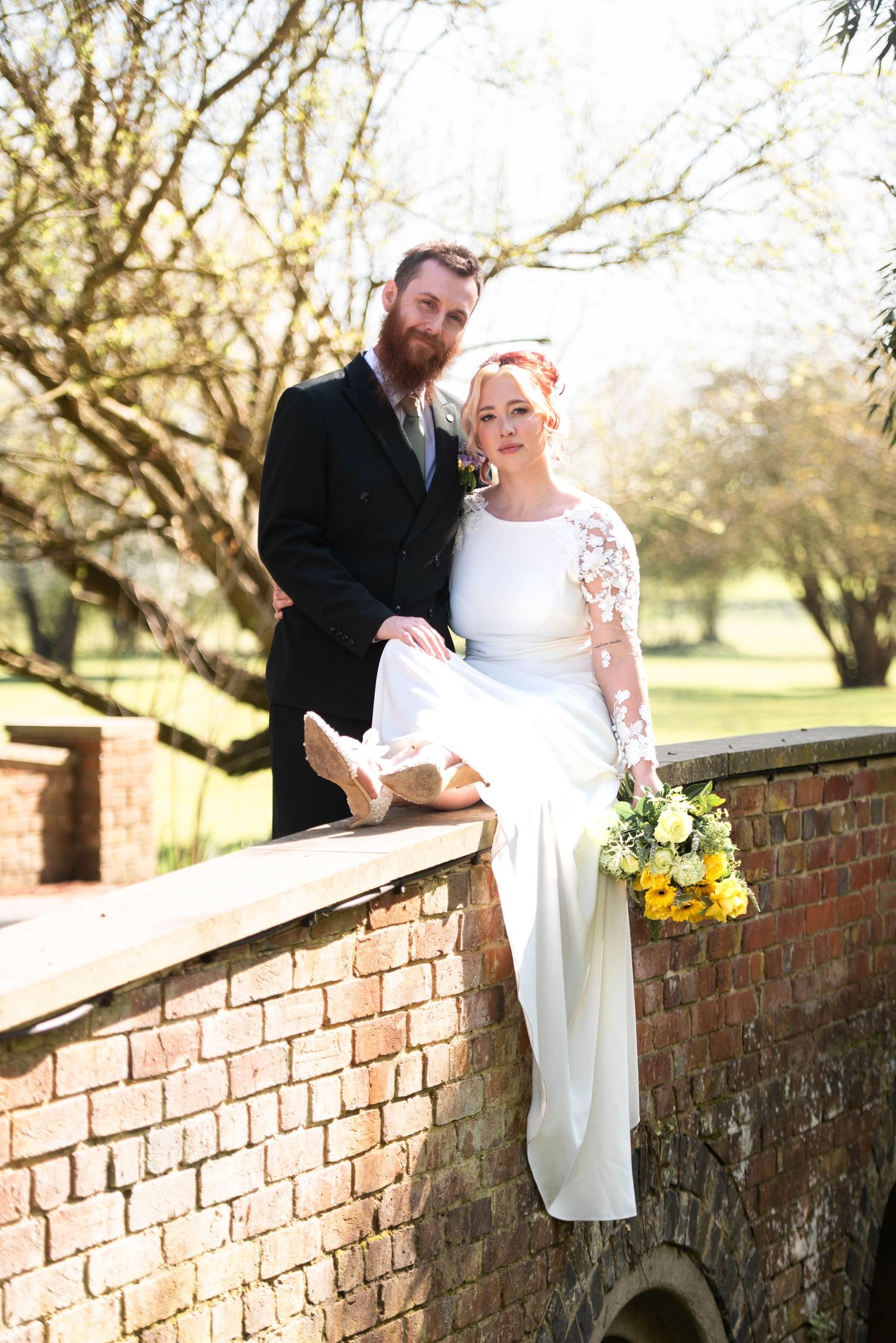 Bride in white lace gown sits on stone bridge wall holding yellow bouquet beside bearded groom in dark suit.