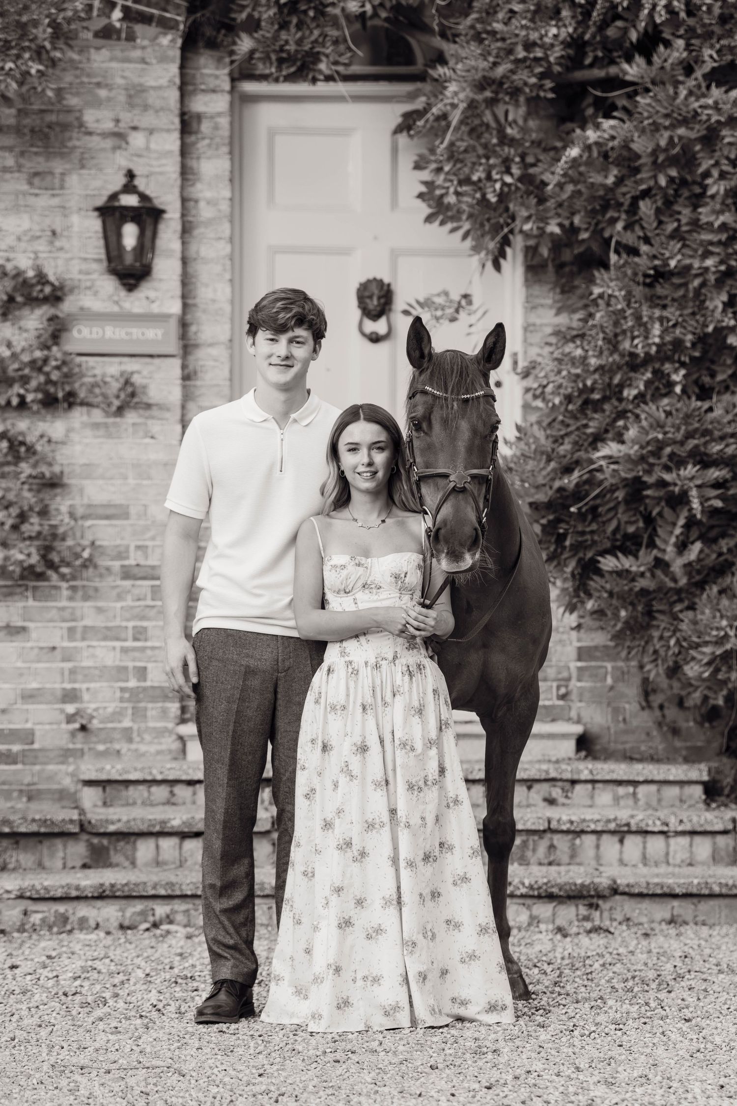 Black and white photo of a young couple posing with a dark horse in front of a brick building.