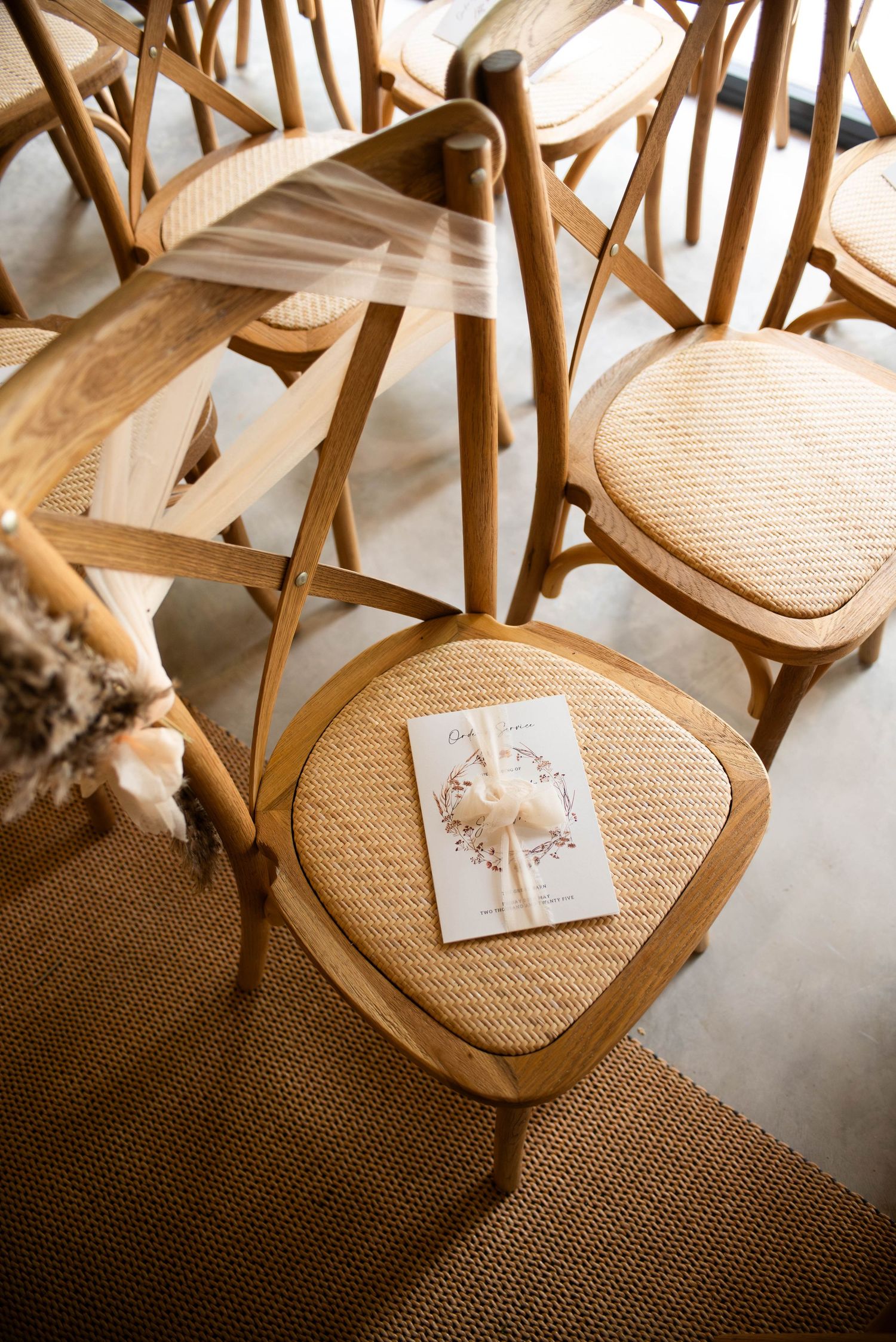 Overhead view of wooden crossback chairs with wedding program on rattan seat cushion.