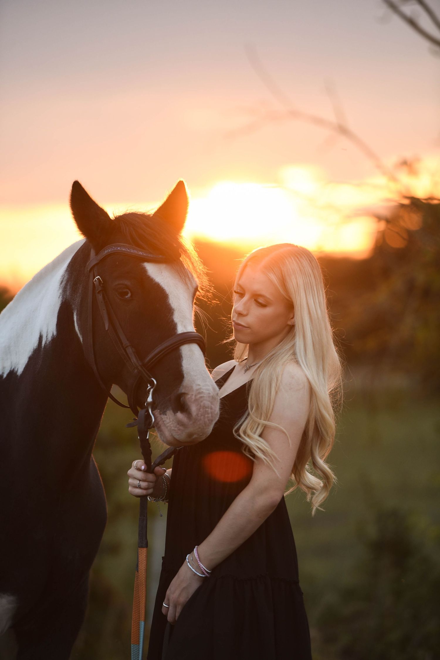 Blonde woman in black dress sharing a tender moment with a paint horse at golden sunset outdoors.