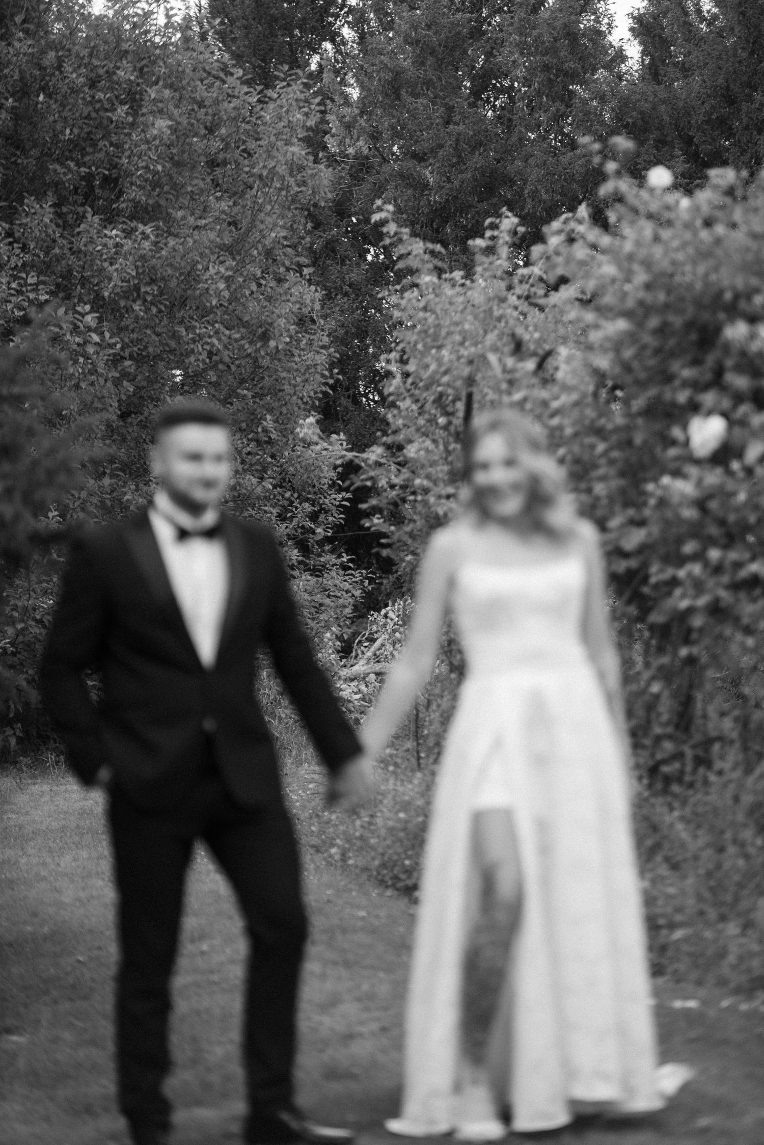 Black and white photo of bride and groom holding hands in a garden, bride wearing a slit wedding gown.