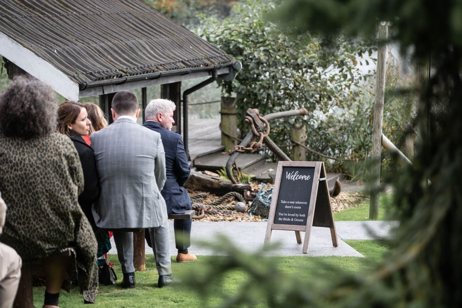Wedding guests mingling outside a rustic venue beside a chalkboard welcome sign on a lawn.