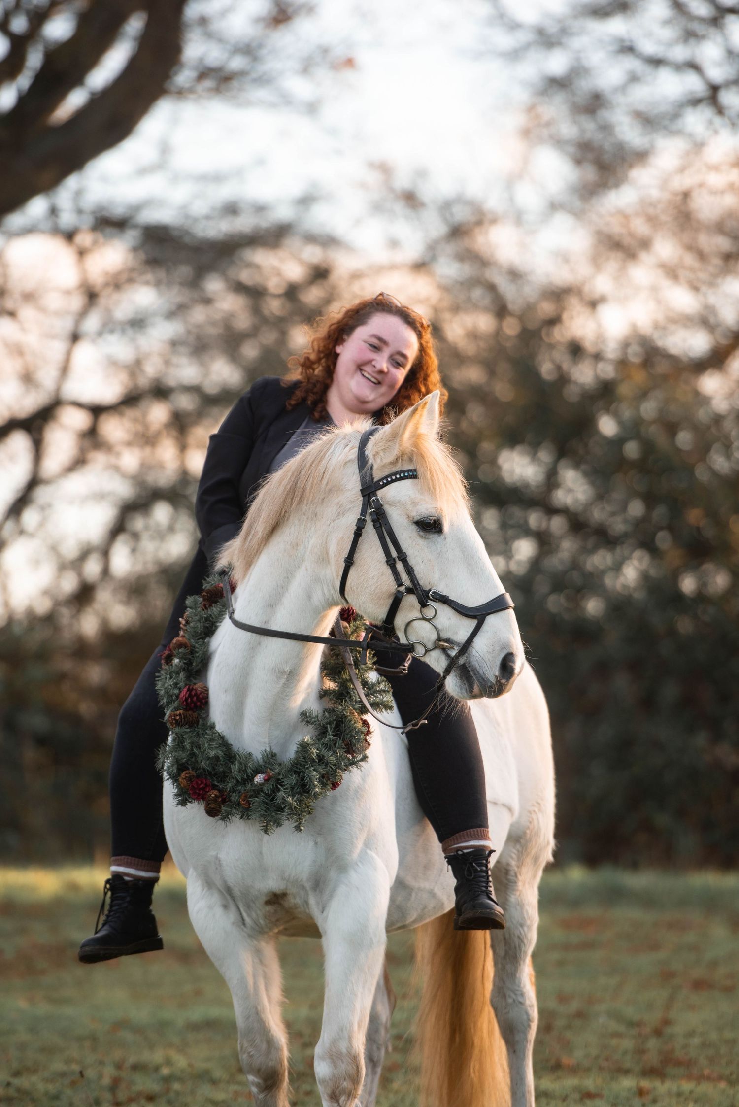 Smiling woman riding a white horse adorned with a holiday wreath in a sunlit outdoor setting.