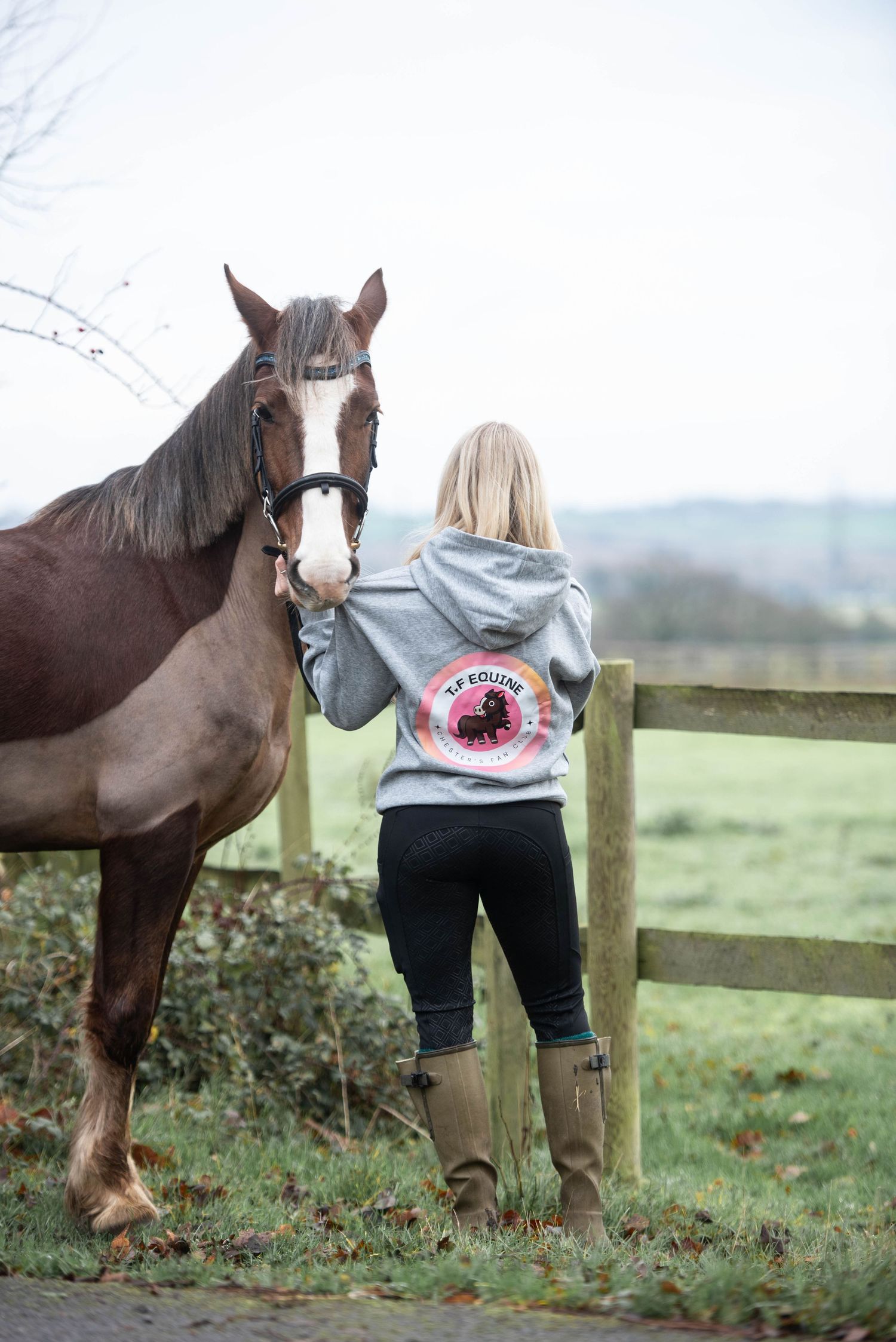 Blonde woman in grey KF Equine hoodie standing with a chestnut horse at a wooden fence outdoors.