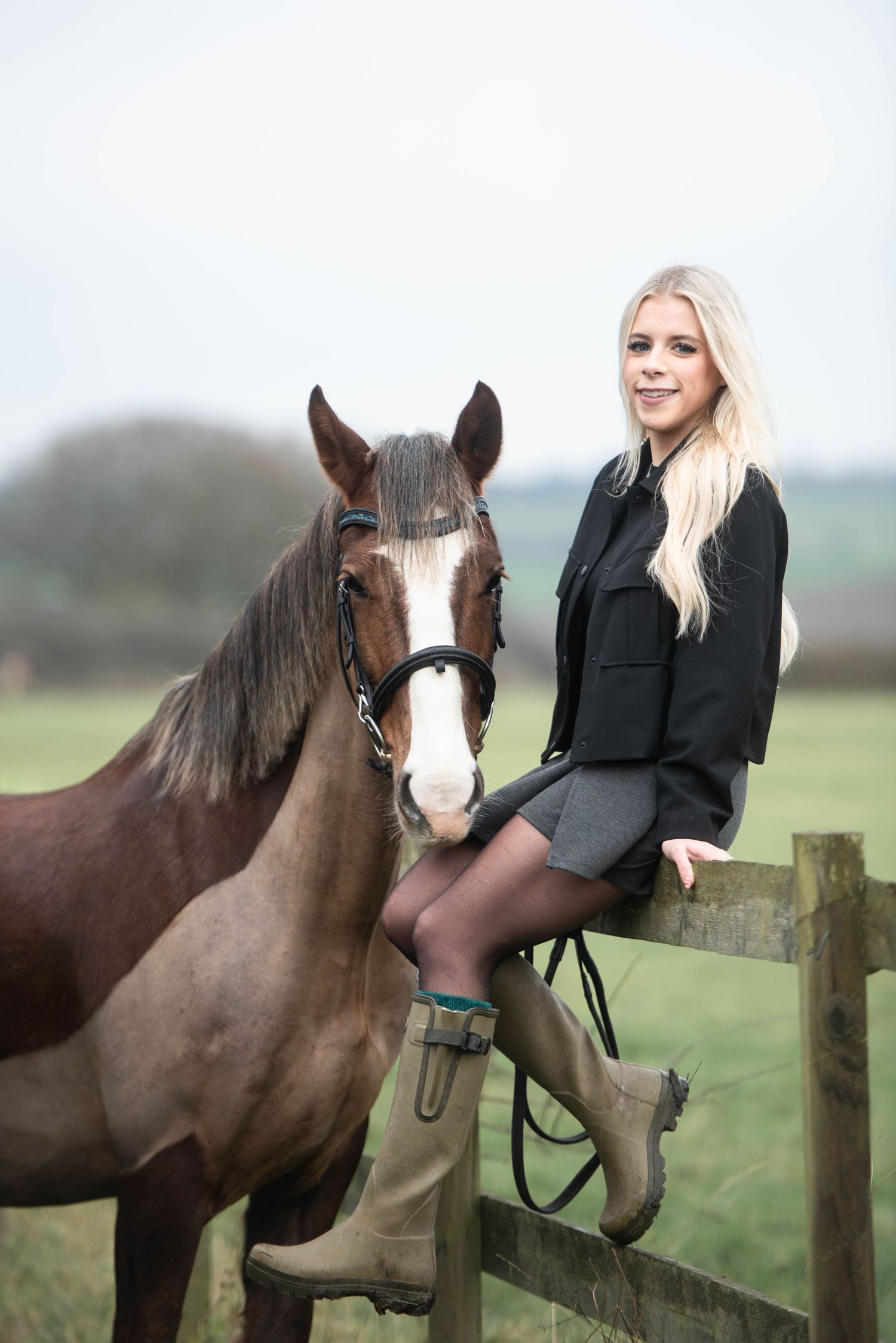 Smiling blonde woman in black jacket sitting on wooden fence beside a chestnut horse in a field.