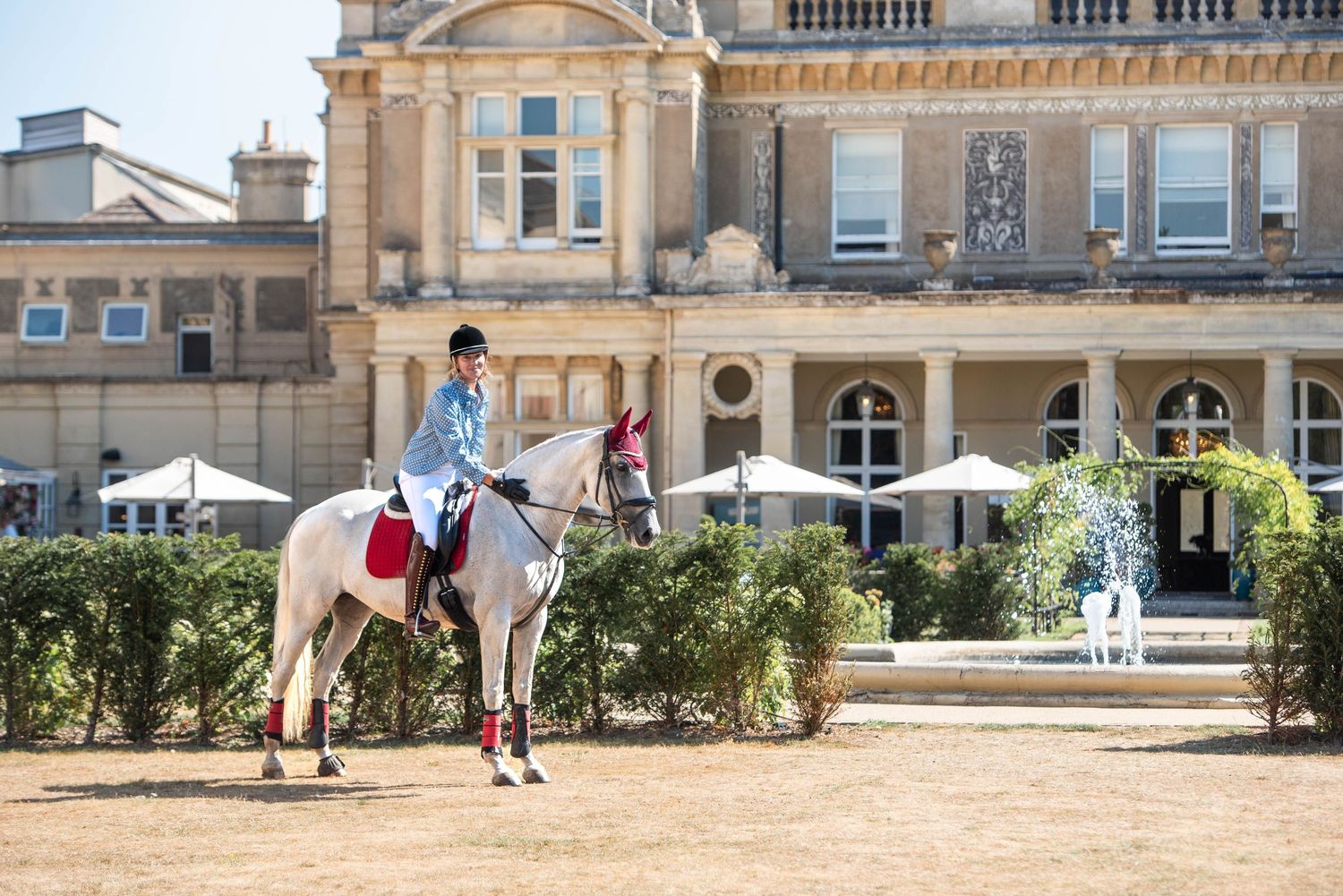 An equestrian rider on a white horse in front of a grand historic mansion on a sunny day.