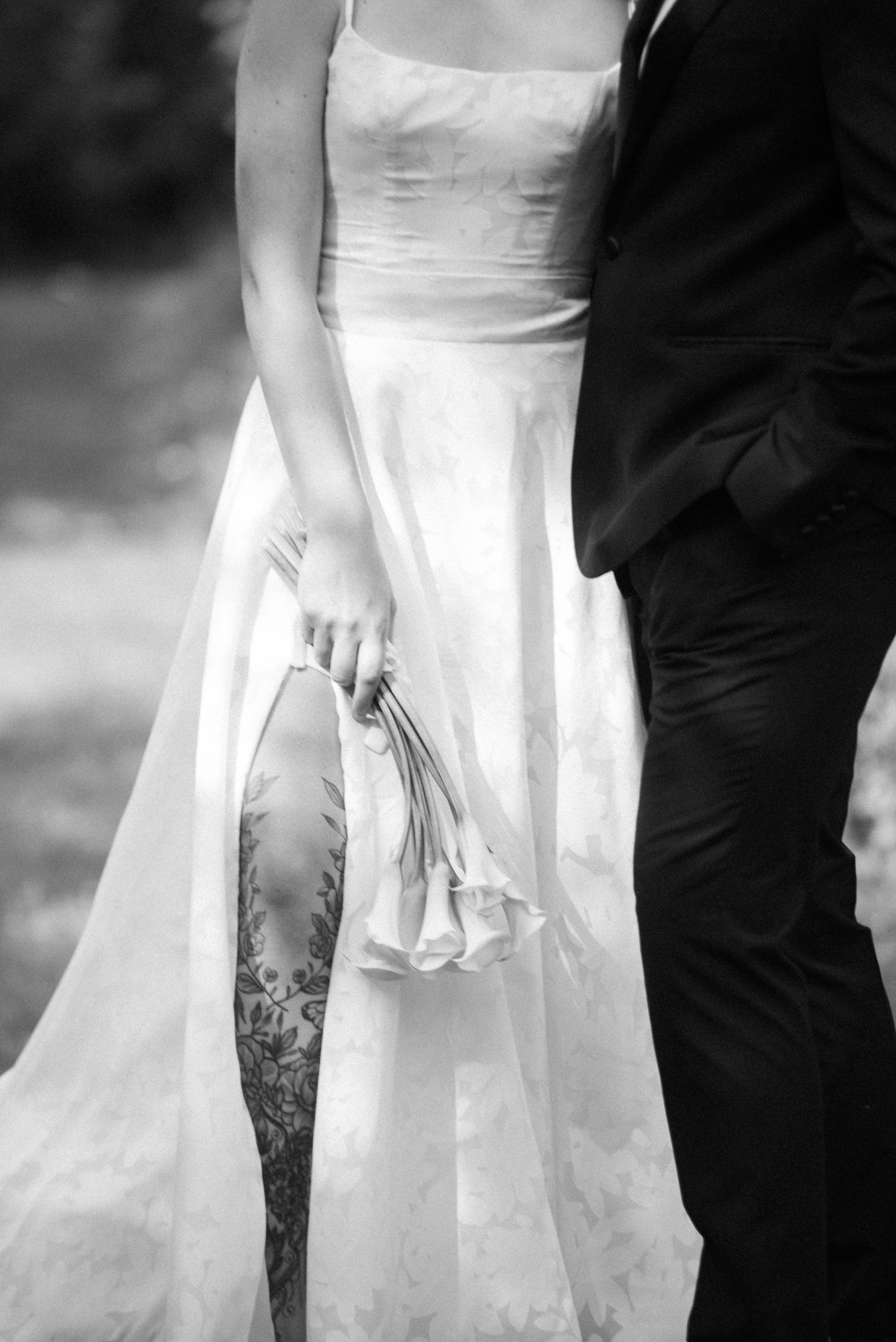 Black and white close-up of bride holding bouquet beside groom, showcasing lace gown and floral tattoo.