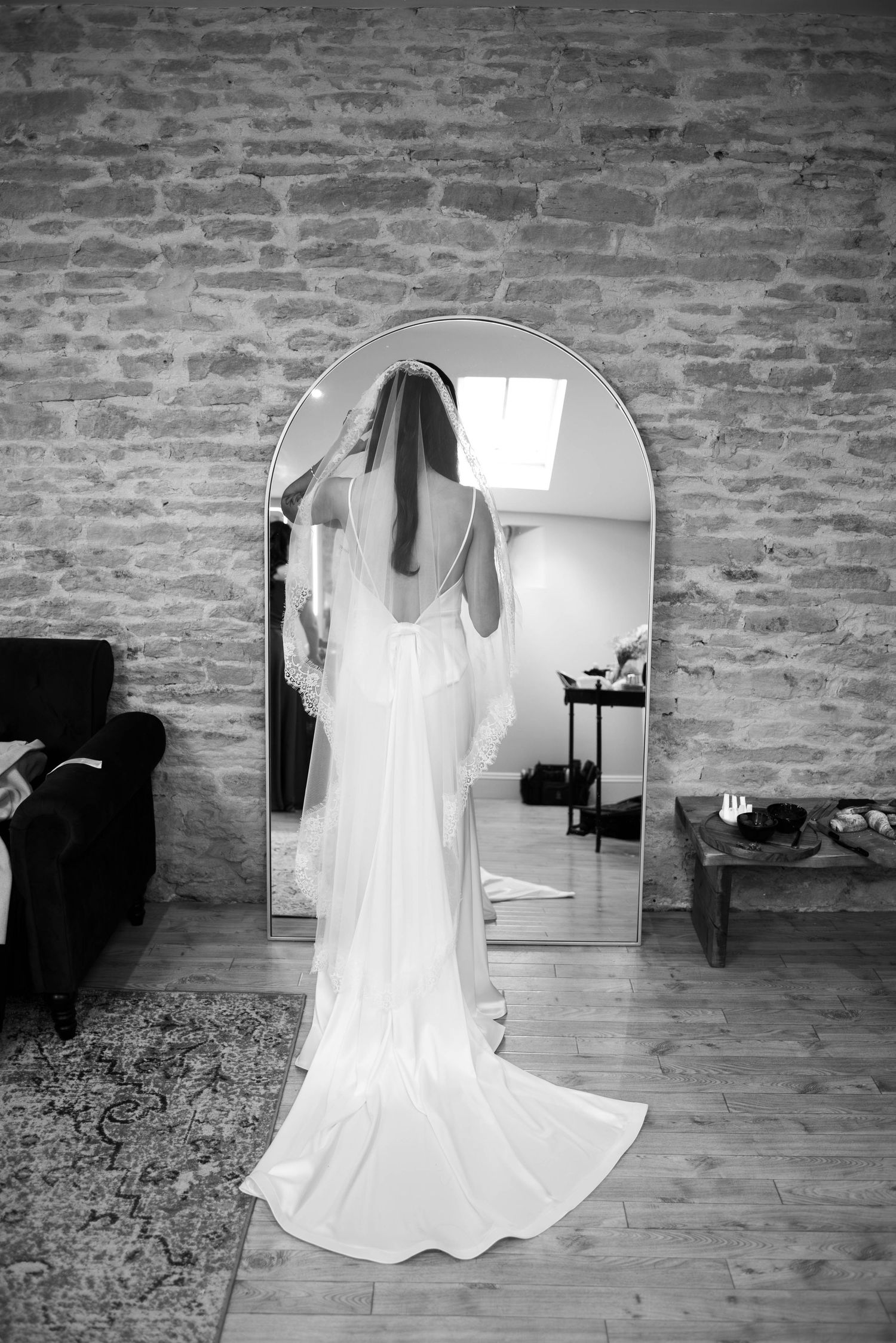 Black and white photo of a bride in a flowing gown and veil reflected in an arched mirror.