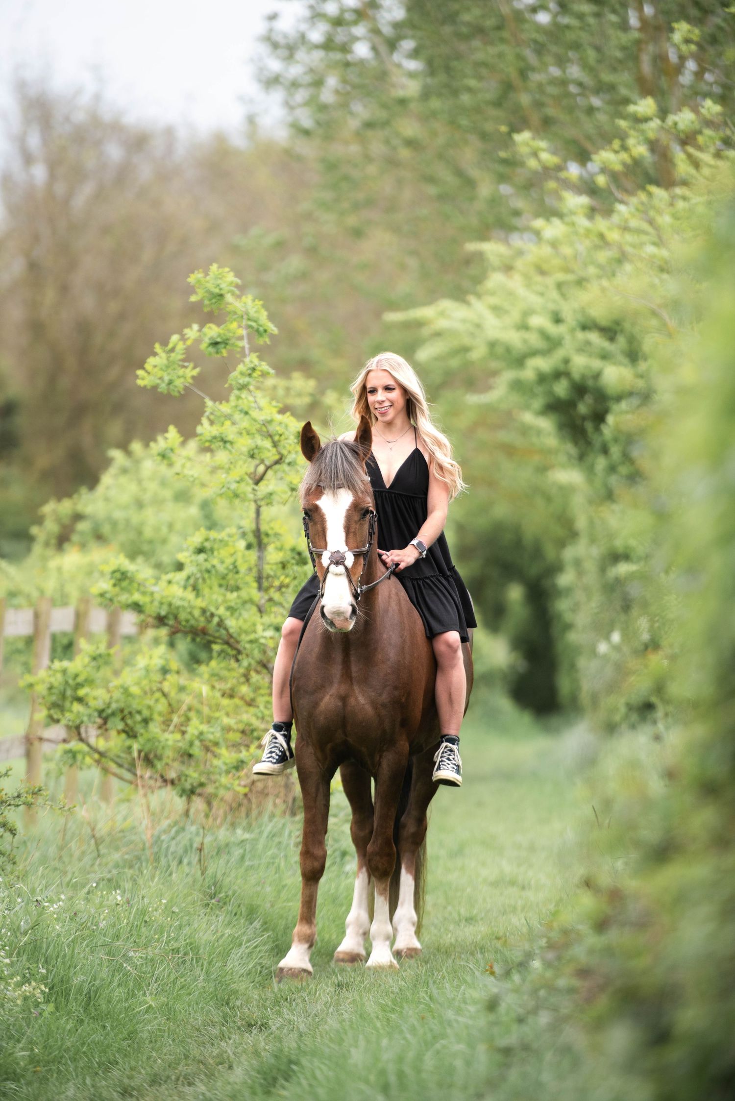 Smiling blonde woman in black dress riding a chestnut horse bareback along a green hedgerow path.