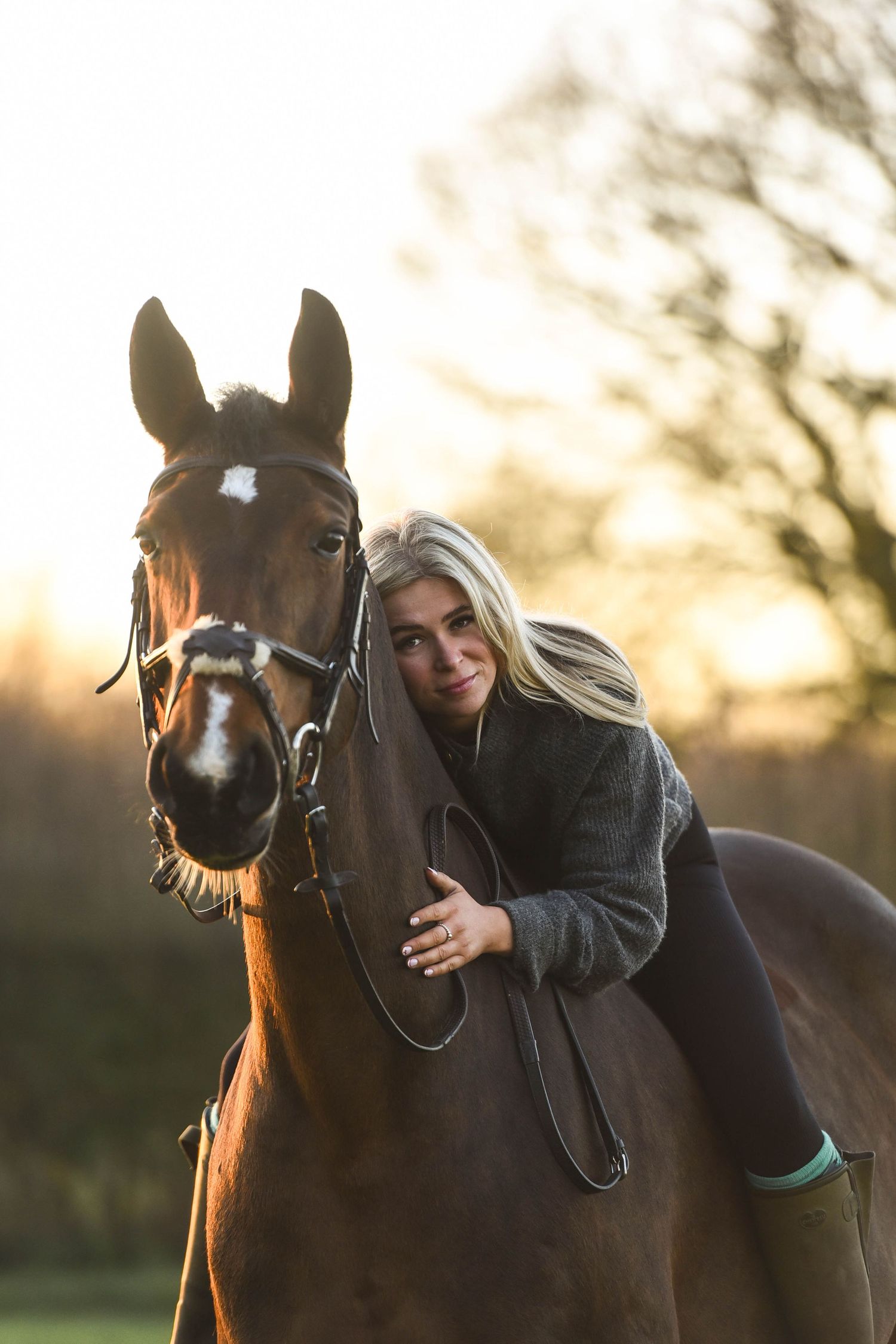 Blonde woman leaning forward hugging a bay horse while riding bareback in warm sunset glow.