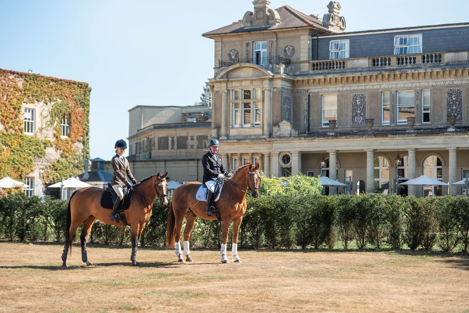 Two riders on chestnut horses outside a grand historic manor house on a sunny day.