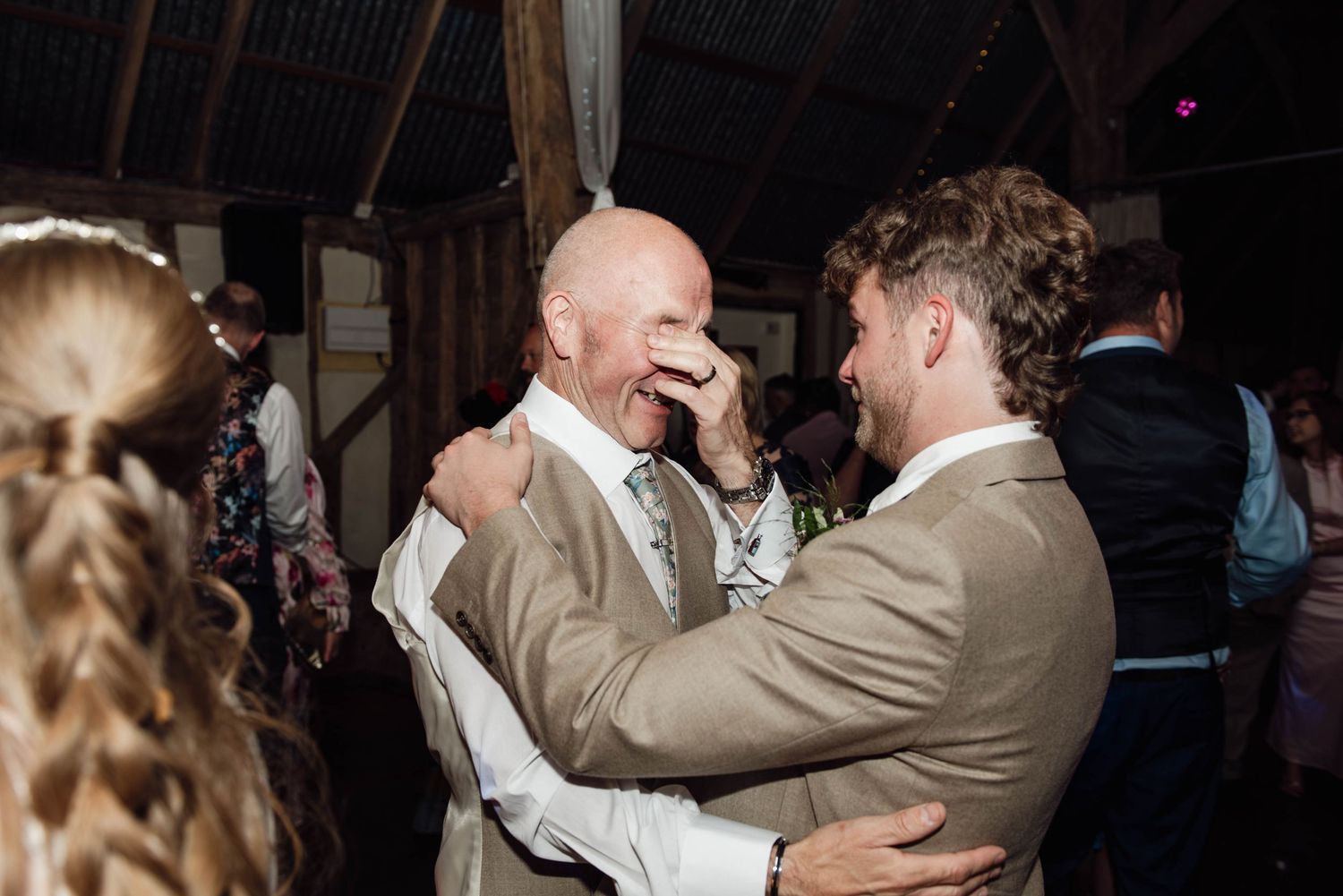 Two men share an emotional embrace on a wedding dance floor in a rustic barn venue.