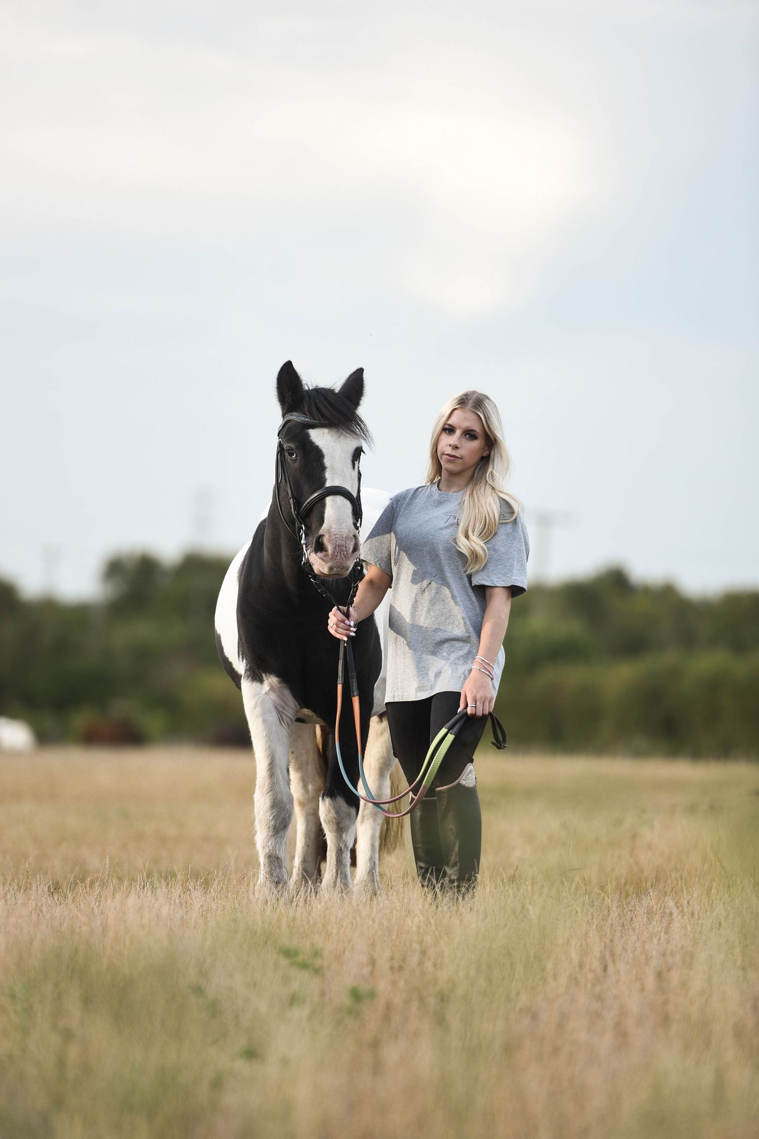 Blonde woman in grey shirt leading black and white paint horse in field.