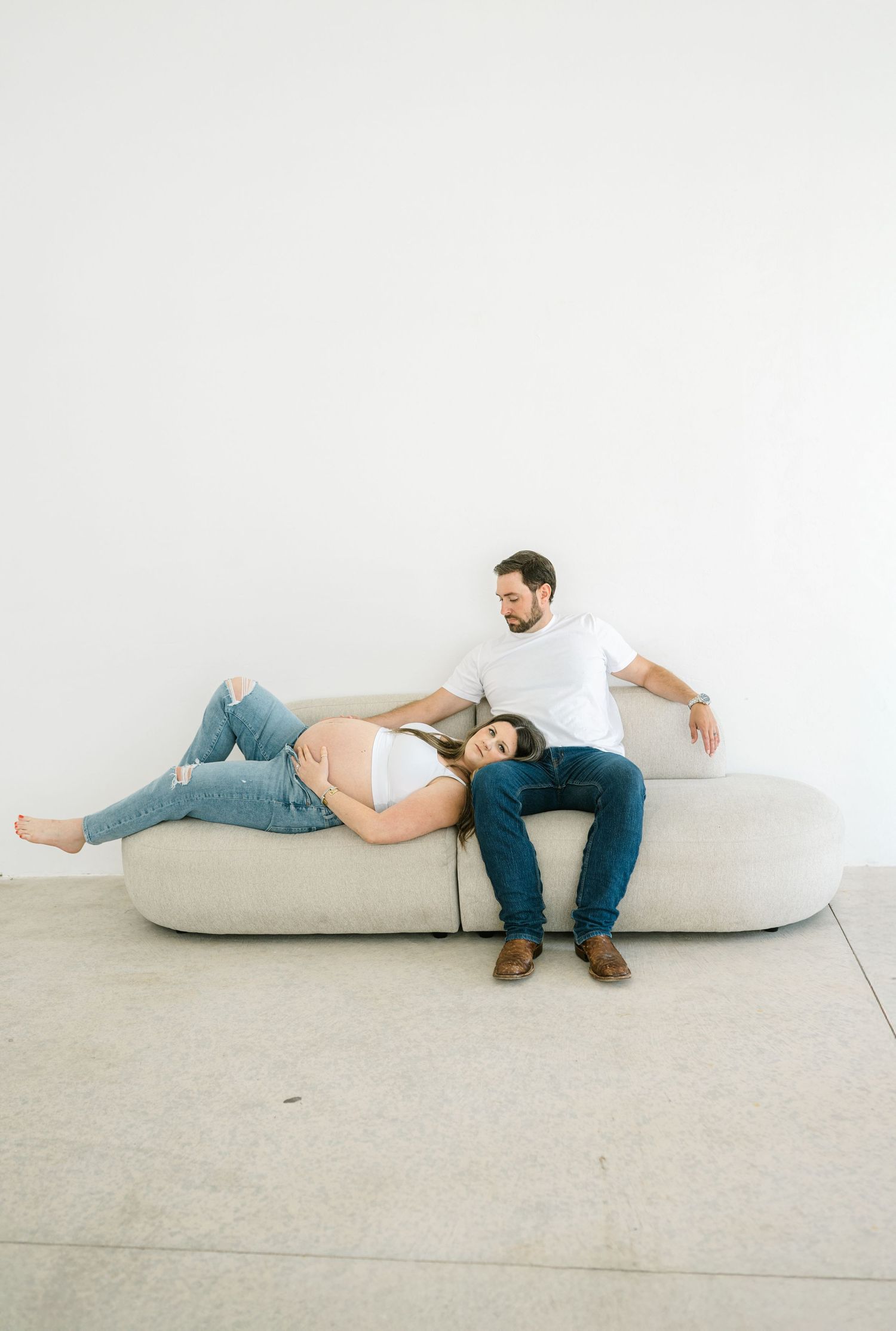 A maternity couple rests together on a minimalist beige couch against a white wall for Oklahoma City maternity photographer