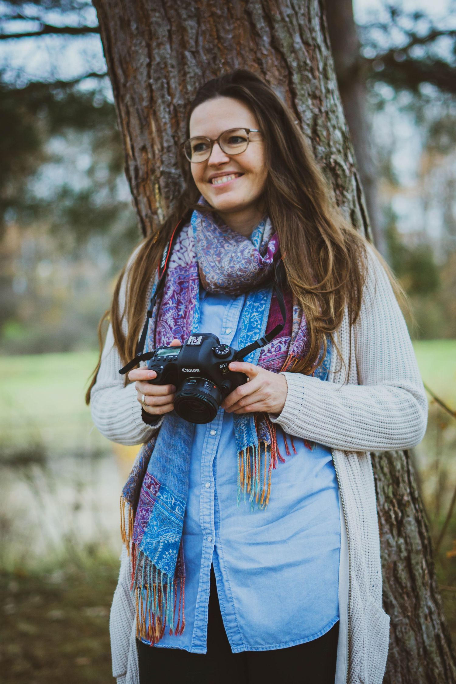 Ein Bild der Fotografin Hanna Witt, die mit Schal und Strickjacke an einen Baum gelehnt im Freien steht und eine Kamera in den Händen hält.