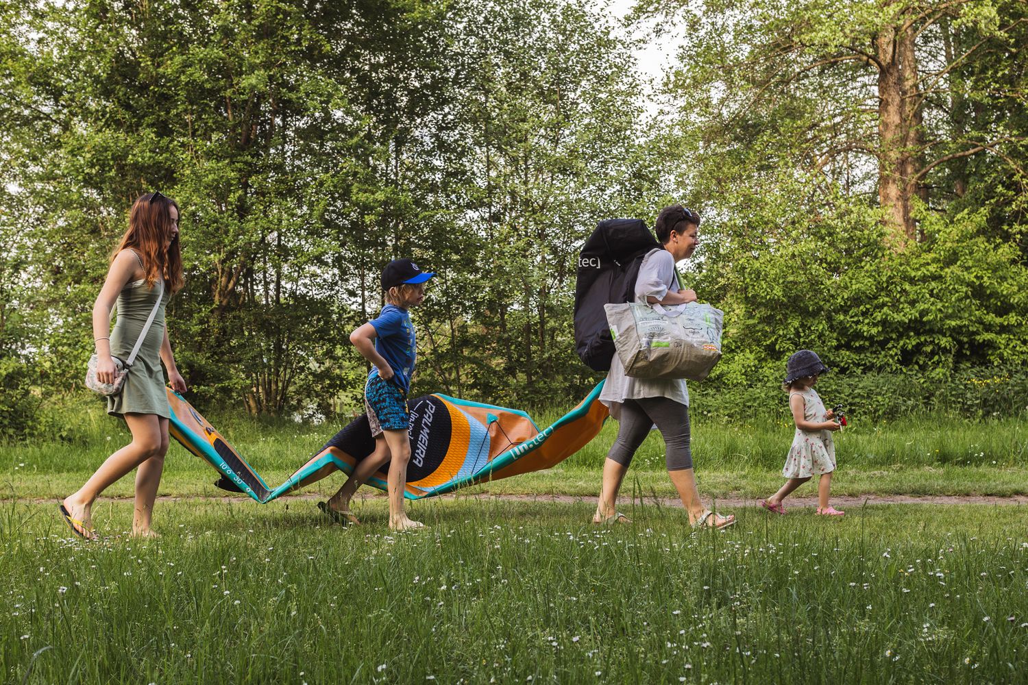 Familienfoto bei dem eine Mutter mit ihren Kindern zu sehen ist die nach einem Ausflug zum Badesee mit einem SUP zurück zum Parkplatz gehen.