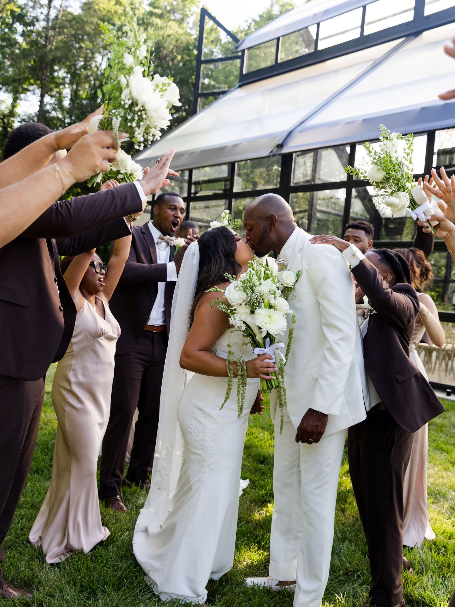 Newlyweds share a kiss surrounded by cheering wedding party in brown and champagne attire on a green lawn.