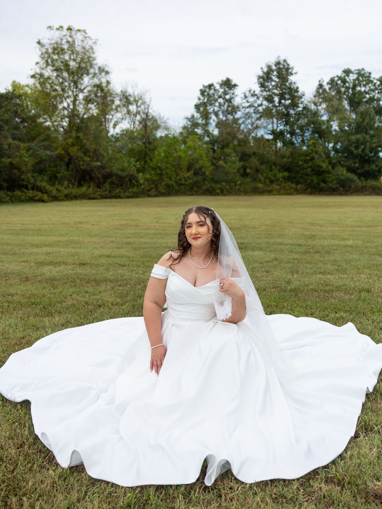 Bride in white off-shoulder ball gown with veil sits gracefully on green grass field with trees in background.