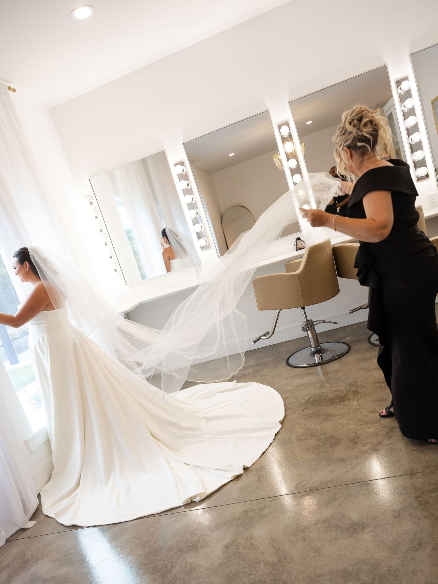 Bride in a white wedding gown with a long veil being arranged by a stylist in a modern bridal salon.