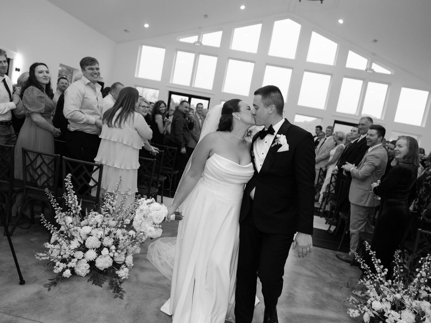 Bride and groom share first kiss at wedding ceremony altar surrounded by guests in elegant white chapel.