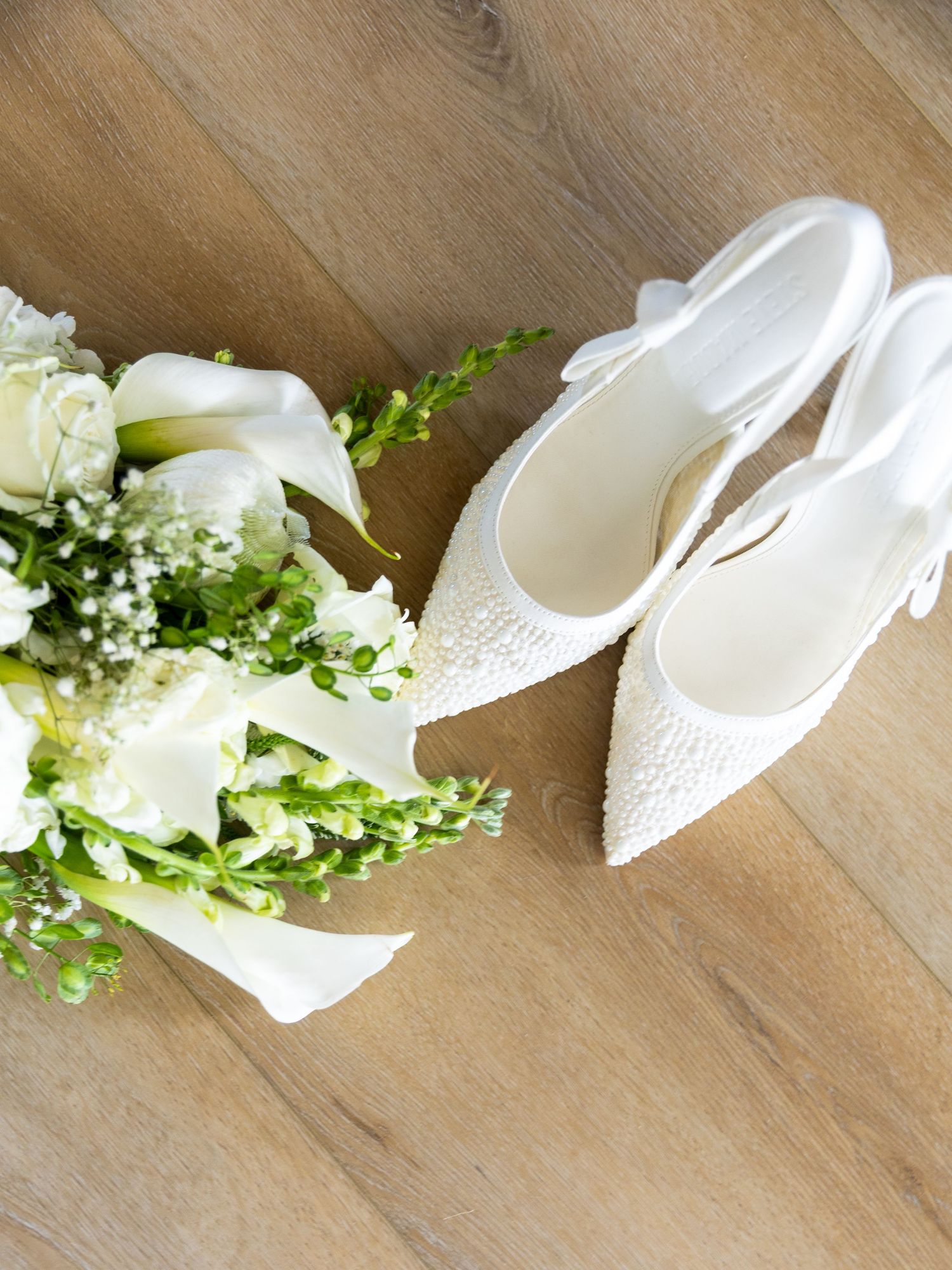 White pearl bridal heels with bow straps beside a white wedding bouquet of roses and calla lilies on wood floor.