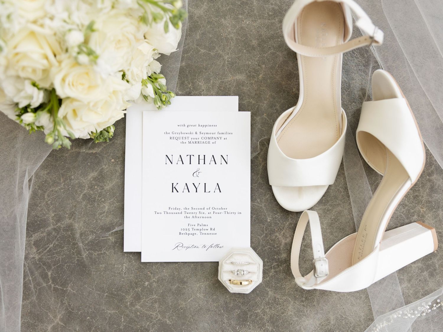 Elegant wedding flat lay with white heels, floral bouquet, ring box, and Nathan & Kayla invitation on marble surface.