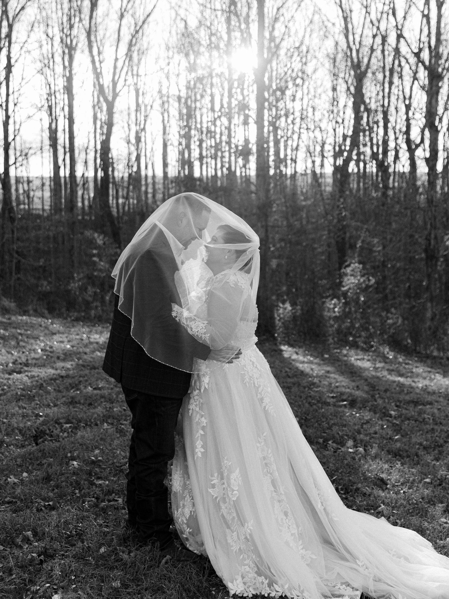 Black and white photo of bride and groom kissing under veil in wooded outdoor setting with sunlight.