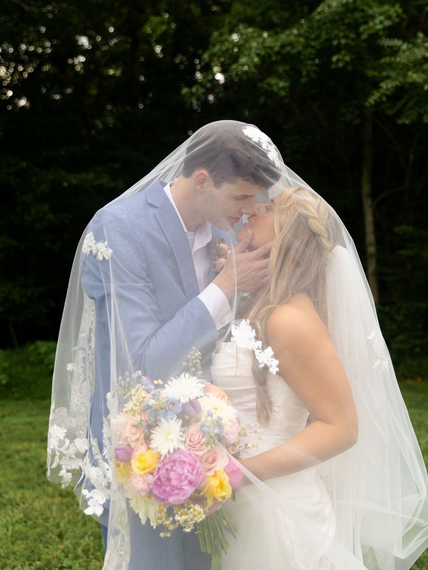 Bride and groom sharing a romantic kiss under a floral wedding veil, bride holding colorful bouquet outdoors.