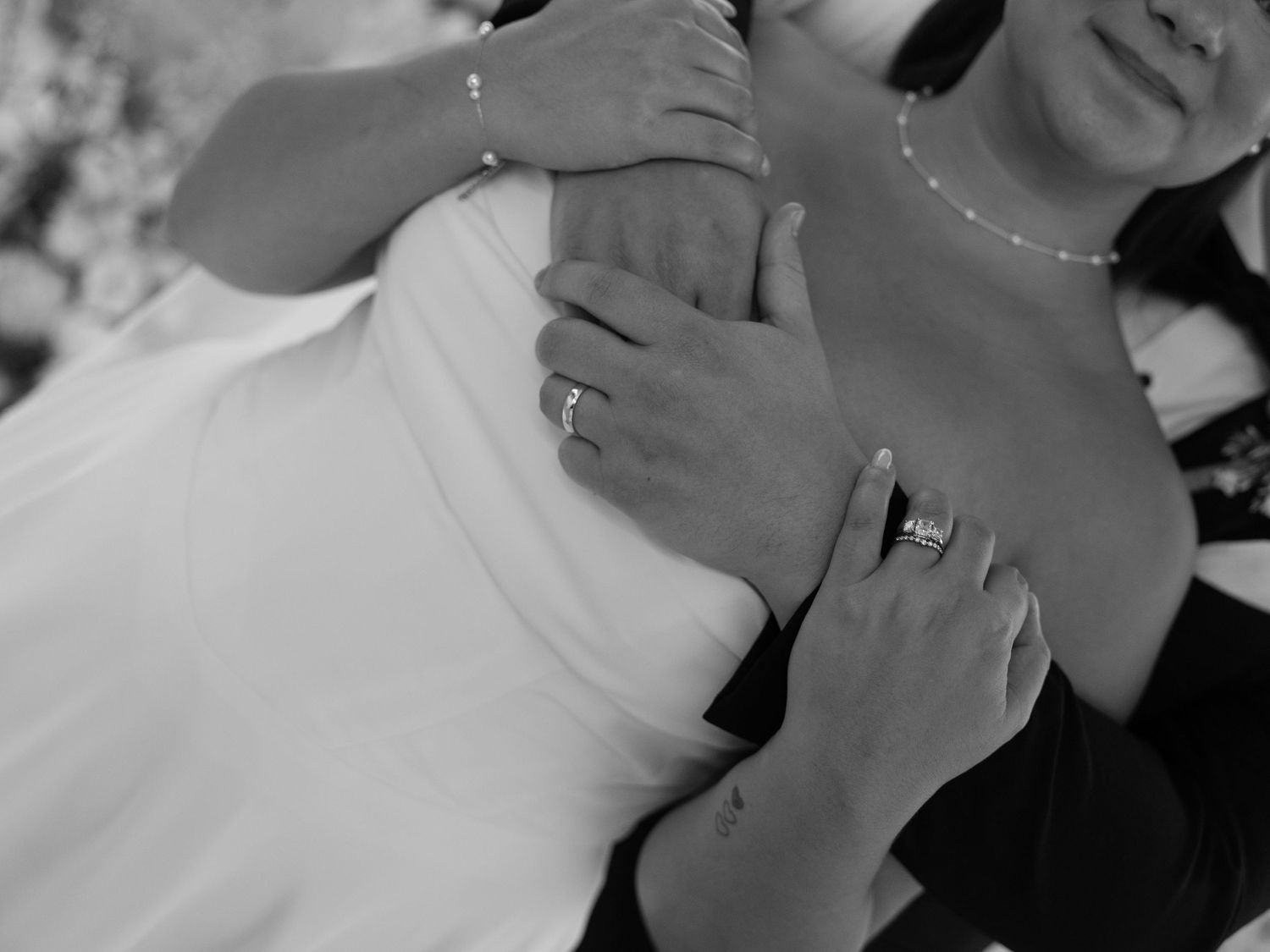 Black and white photo of a bride in a white dress being embraced, showing wedding rings on hands.