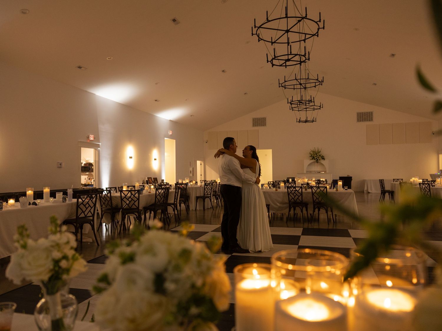 Couple shares first dance in elegant candlelit wedding reception hall with chandeliers and white floral arrangements.